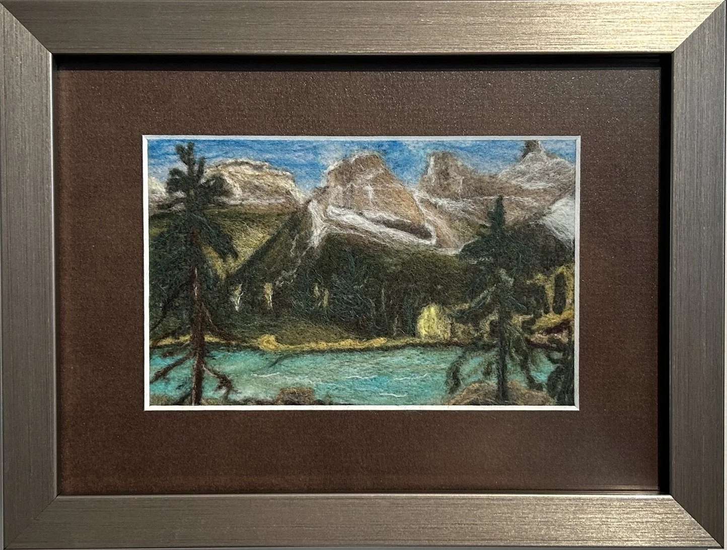 A Needle felted landscape featuring a mountain range, pine trees, and a body of water in the foreground, with a blue sky.