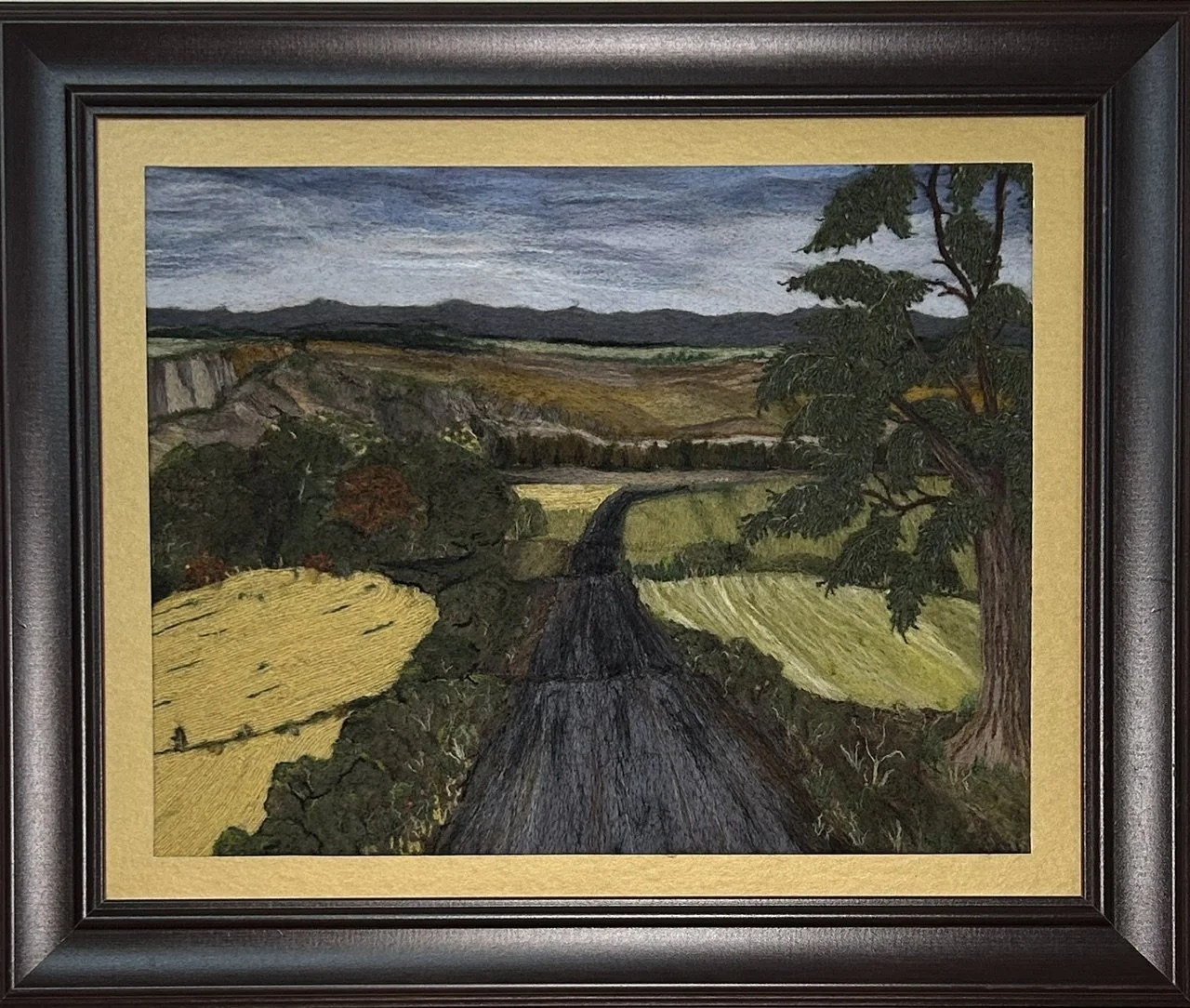 A needle felted landscape depicting rolling wheat fields and green meadows with a road flowing through the center, mountains in the distance and a large spruce tree on the right, under a mixed sky.