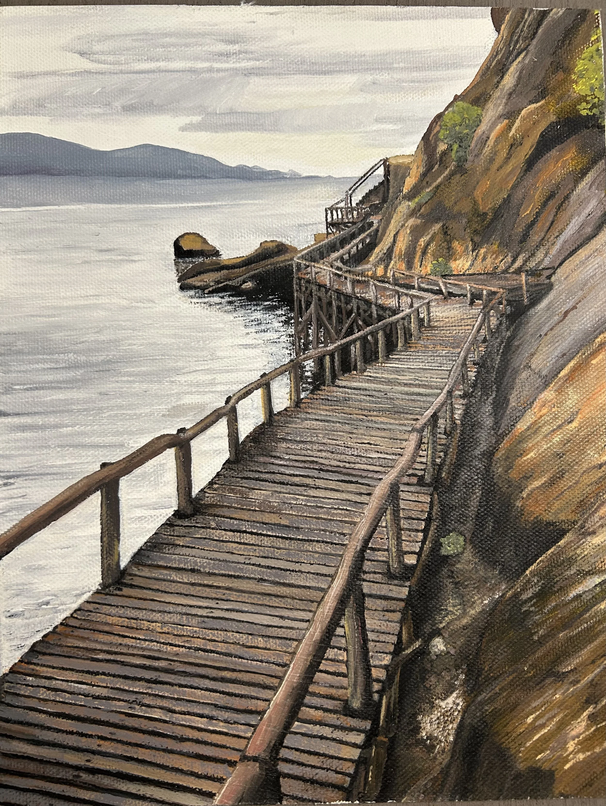 A wooden walkway along a rocky coastal cliff with a view of water and distant mountains under a cloudy sky.