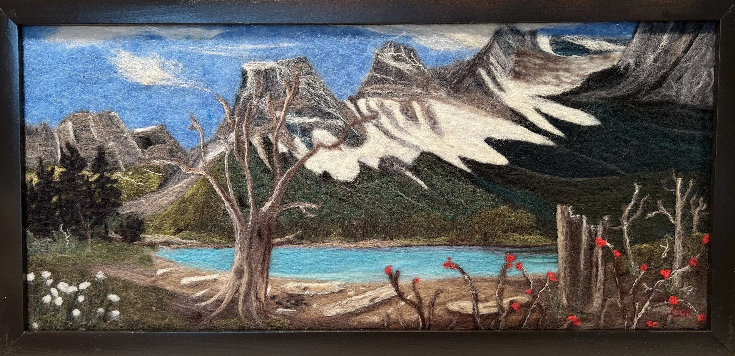 A landscape scene painted with felt depicting mountains, a blue lake, trees, and clouds.