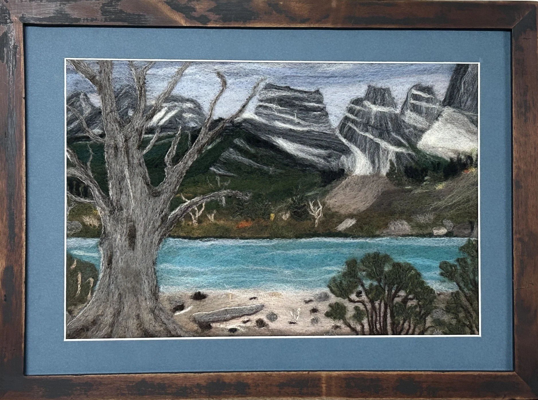 A felted landscape of 3 Sisters mountains with a blue lake, and a weathered old tree on the beach in the foreground.
