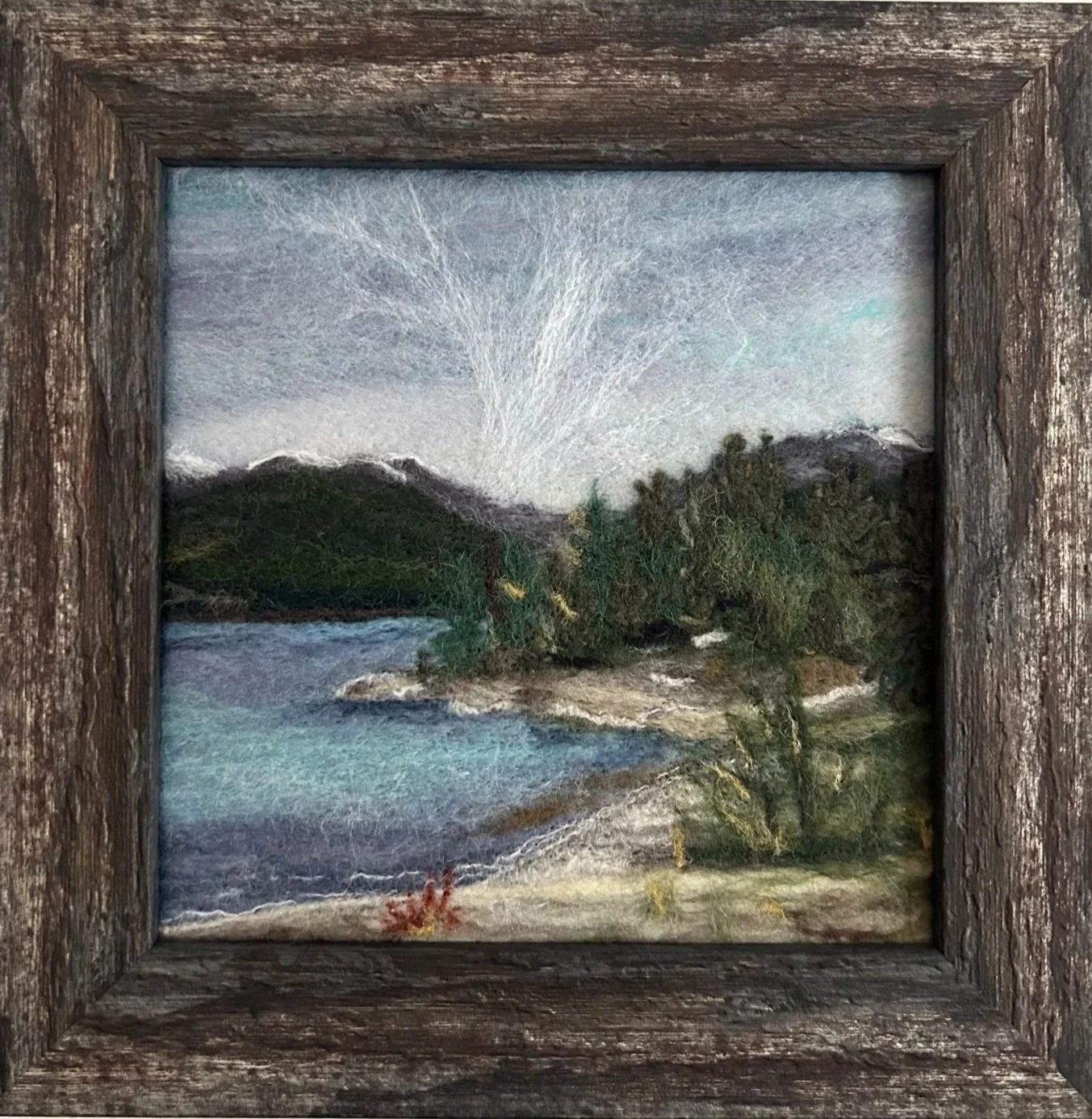 Needle felted landscape of a lake with green hills, a sandy beach, and a fanning cloud in the blue sky.