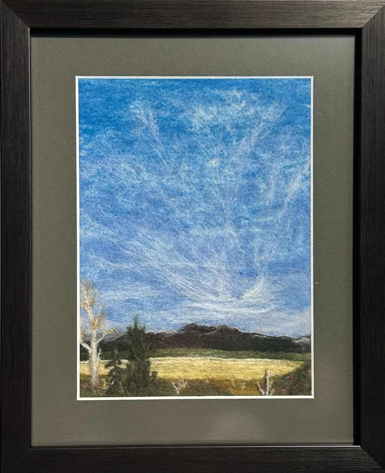 Needle felted landscape of a bright blue sky with wispy exploding white clouds and a rural scene with wheat fields, mountains. and trees in foreground