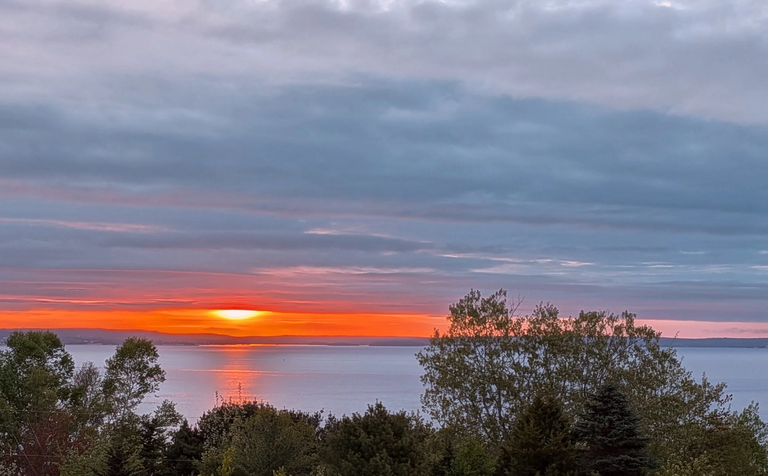 Sunrise over the water in Nova Scotia, a band of orange light beneath grey cloud, with trees in the foreground.