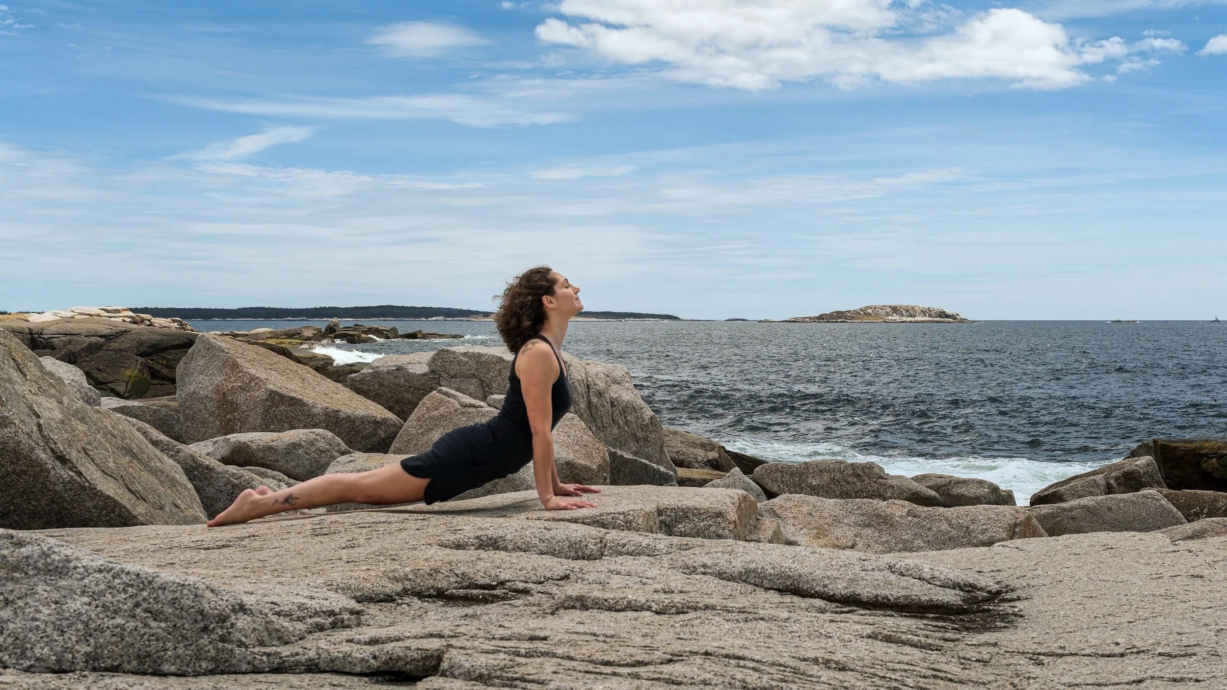 A woman practicing yoga on a rocky shoreline by the ocean, performing upward dog pose with her hands and feet on the rocks, looking upward, with a cloudy sky and distant islands in the background.