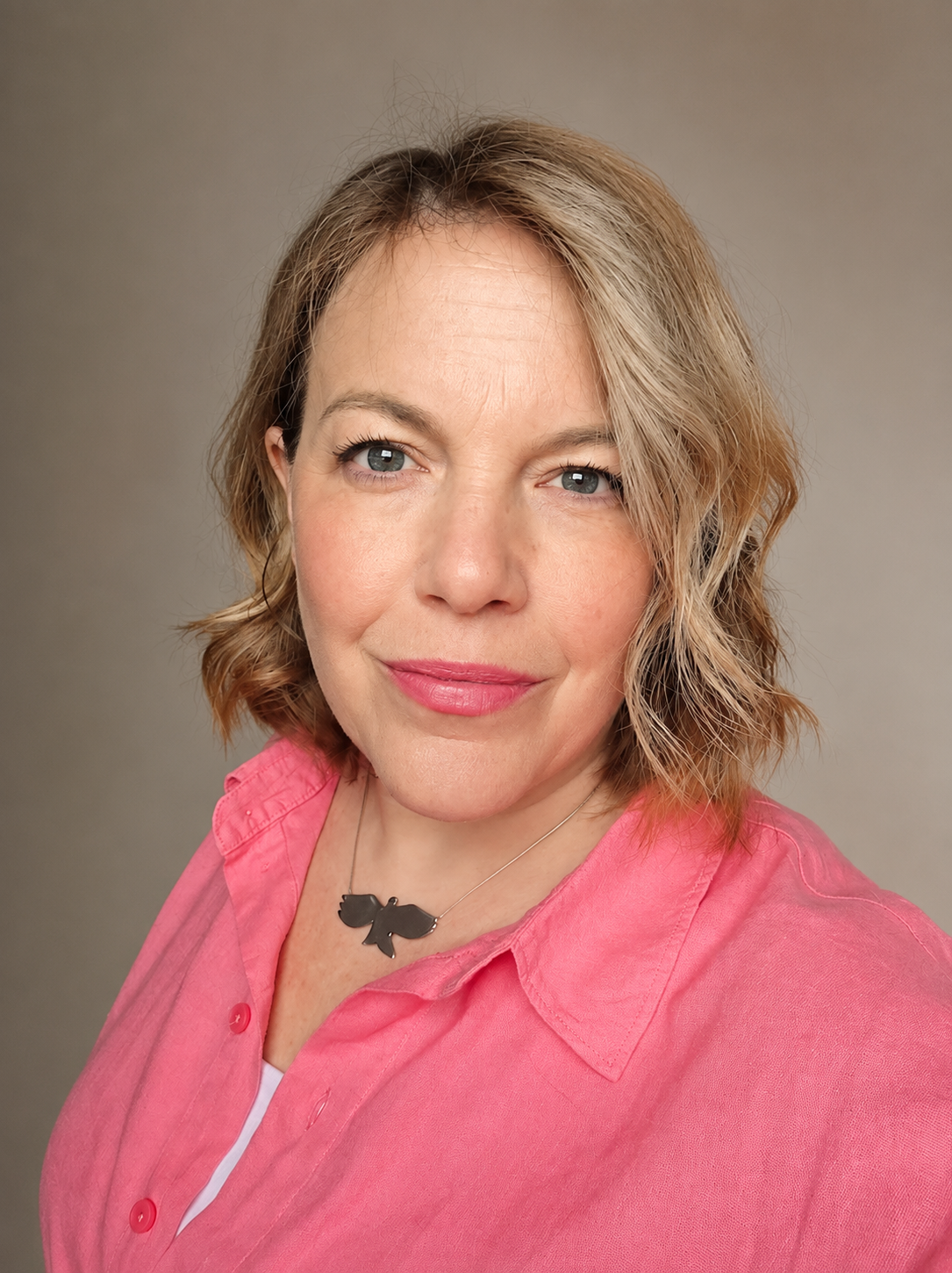 A woman with shoulder-length wavy blonde hair and blue eyes, smiling, wearing a pink button-up shirt and a black butterfly necklace against a neutral background.