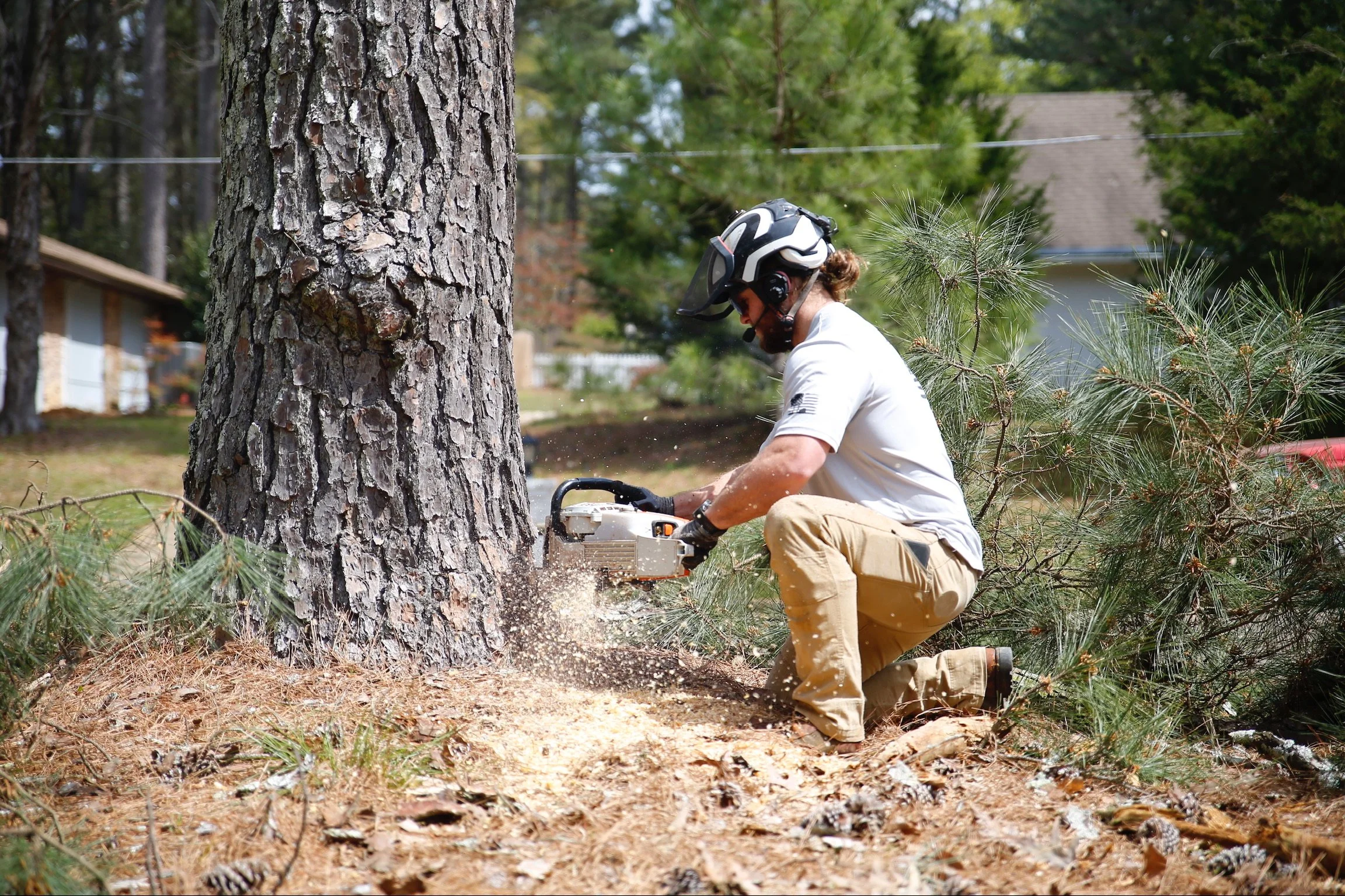 A man with a helmet using a chainsaw to cut down a large tree in a residential yard.