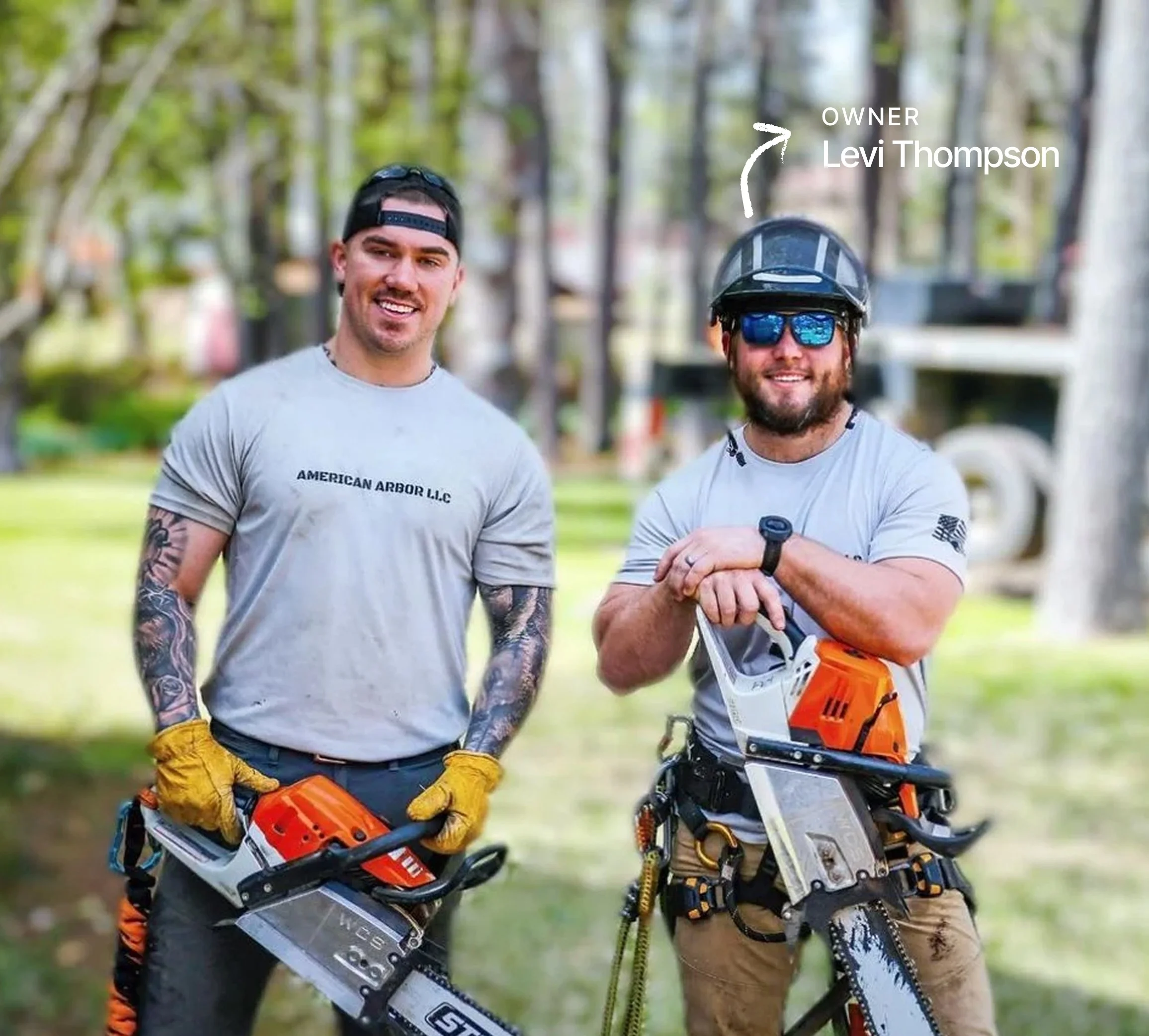 Two smiling men standing outdoors in a park, dressed in casual shirts and safety gear, holding chainsaws, with a background of trees and grass.
