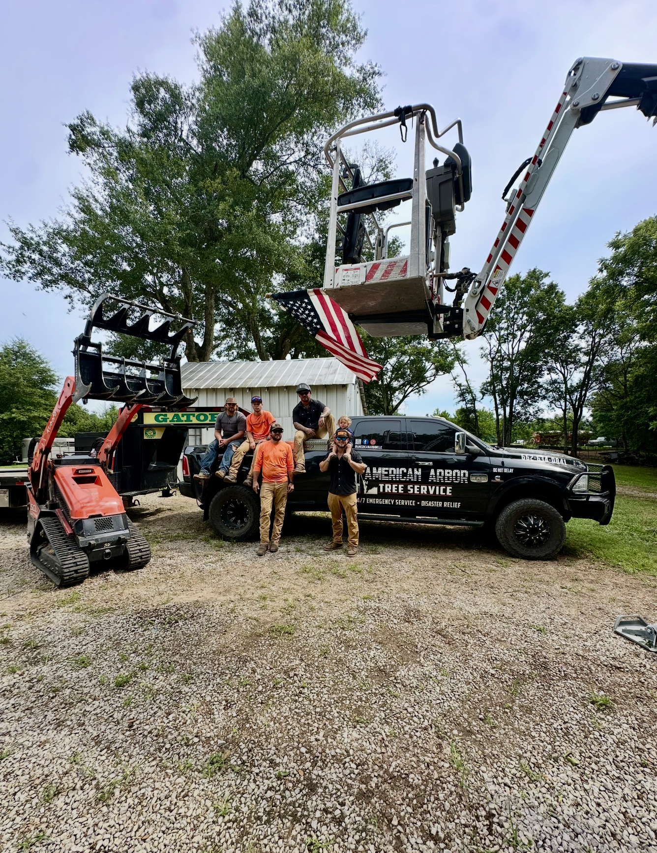 A group of five men standing and sitting on a black pickup truck with 'American Arbor Tree Service' branding parked outdoors on gravel. One person is in an elevated cherry picker basket above the truck, holding an American flag. A small tracked loader is parked beside the group, and a white shed and leafy trees are in the background.