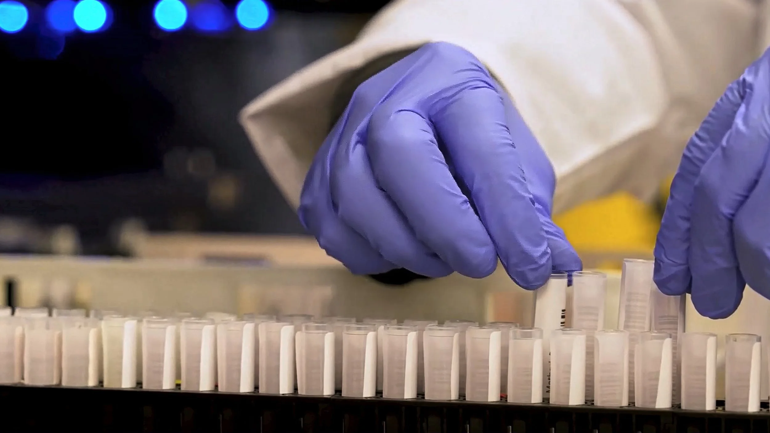 A scientist wearing purple gloves handling test tubes in a laboratory setting, with blue lights in the background.