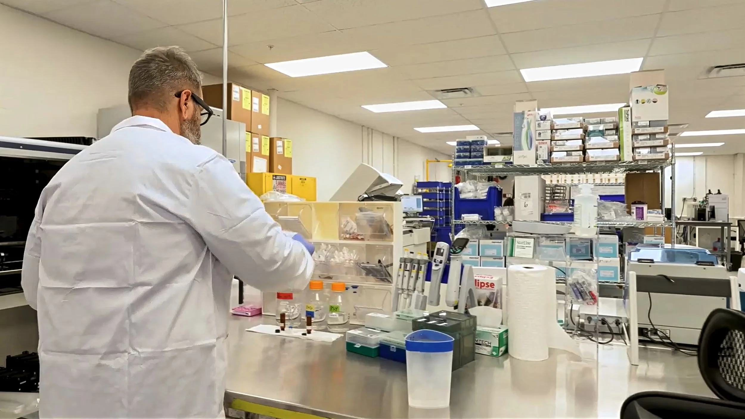 A scientist or technician in a white lab coat working at a lab bench in a scientific laboratory, with shelves of lab supplies and equipment in the background.