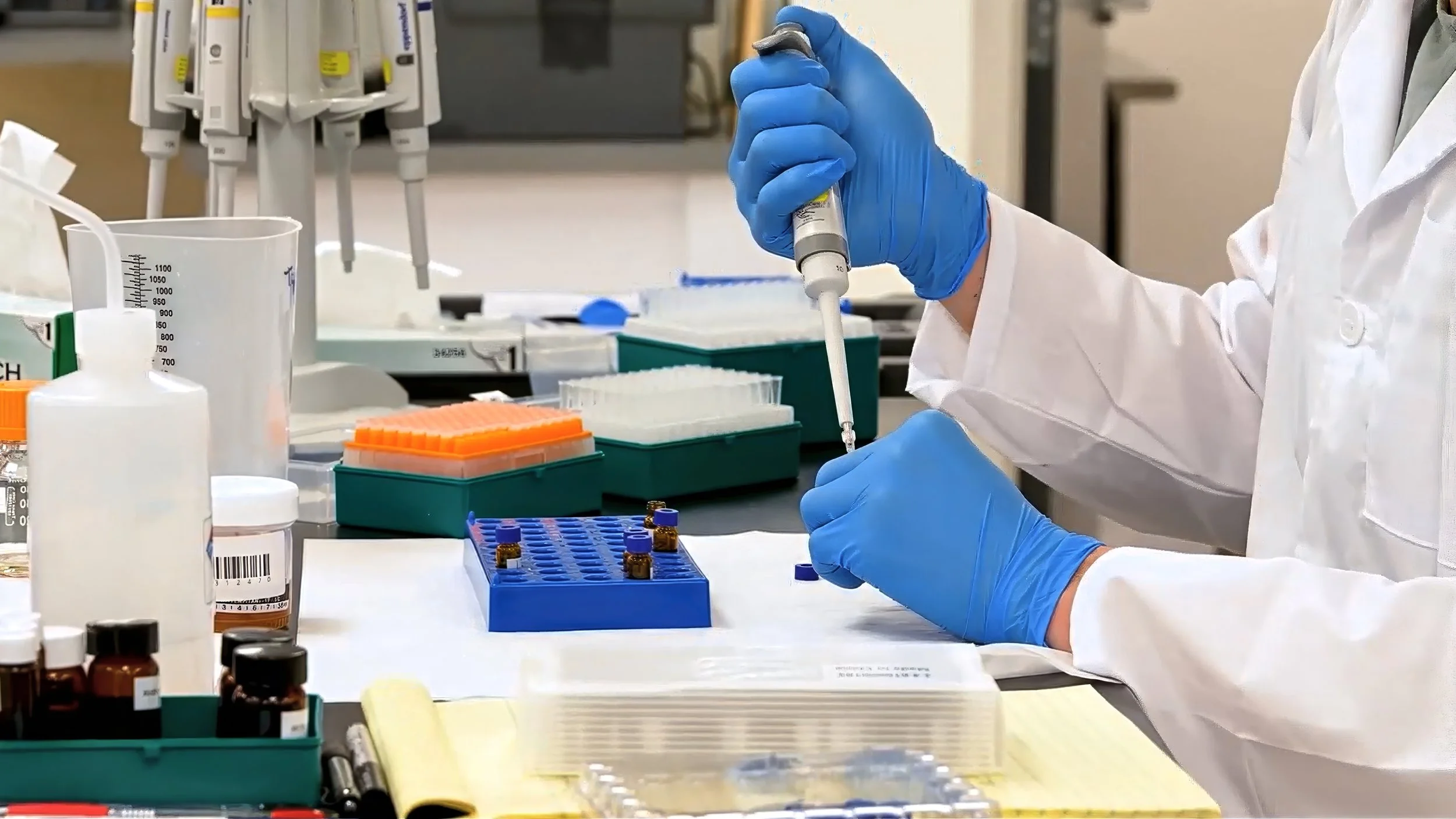 A scientist wearing blue gloves and a white lab coat is using a micropipette to work with small vials and samples in a laboratory setting. Various lab equipment and containers are on the workspace.
