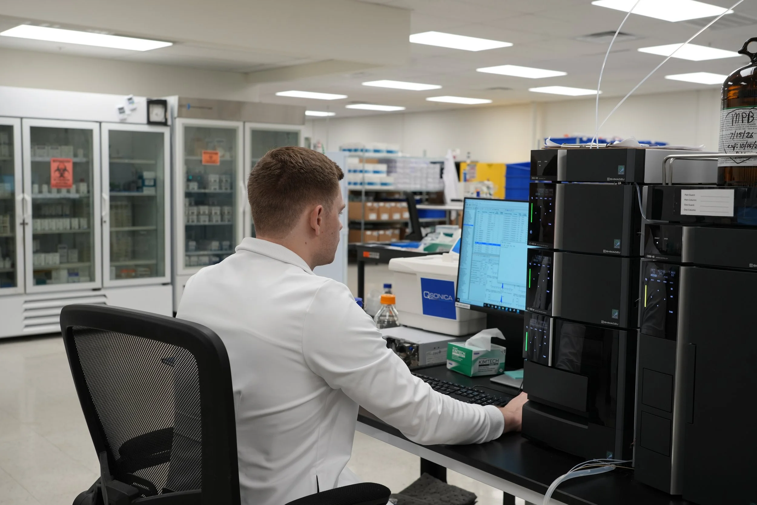 A scientist working in a laboratory, seated at a desk with a computer and laboratory equipment, surrounded by cabinets and shelves filled with supplies.