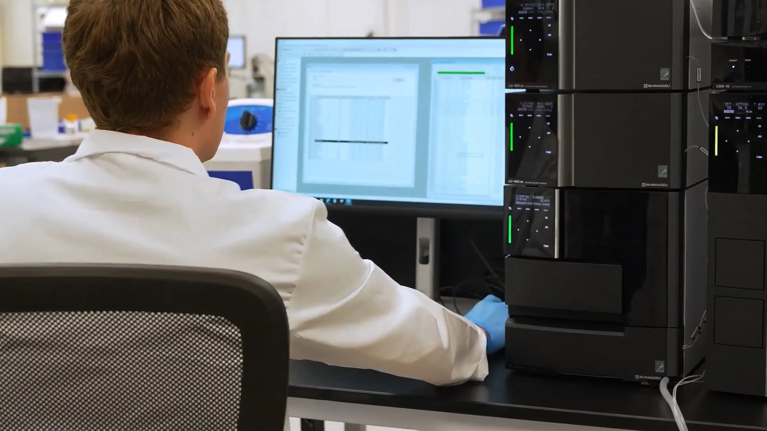 A scientist wearing a white lab coat working at a computer in a laboratory with lab equipment around.