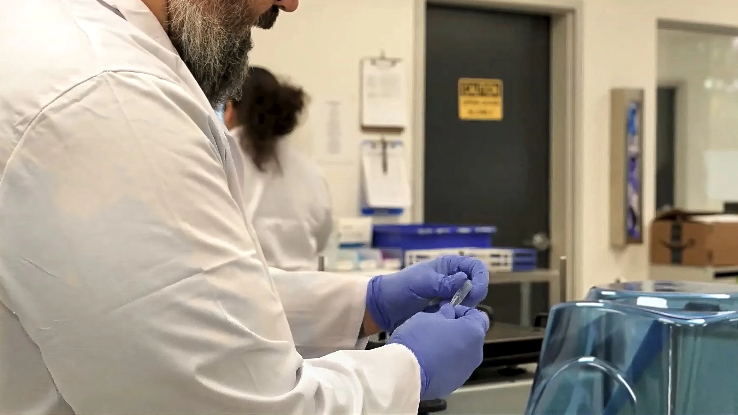 A healthcare worker in a white coat and blue gloves preparing a syringe in a medical setting, with medical supplies and storage containers nearby.