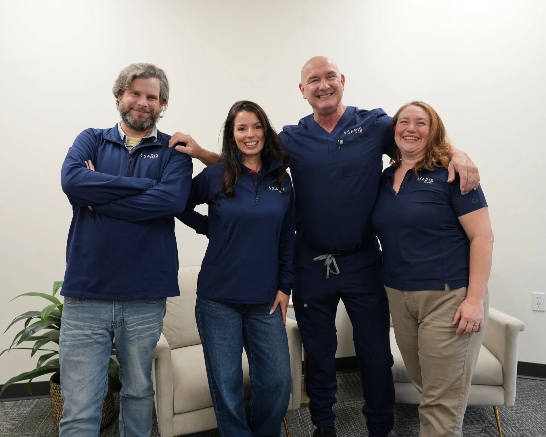 Group of four smiling healthcare professionals standing together in an office, wearing navy blue scrubs with a white wall background.