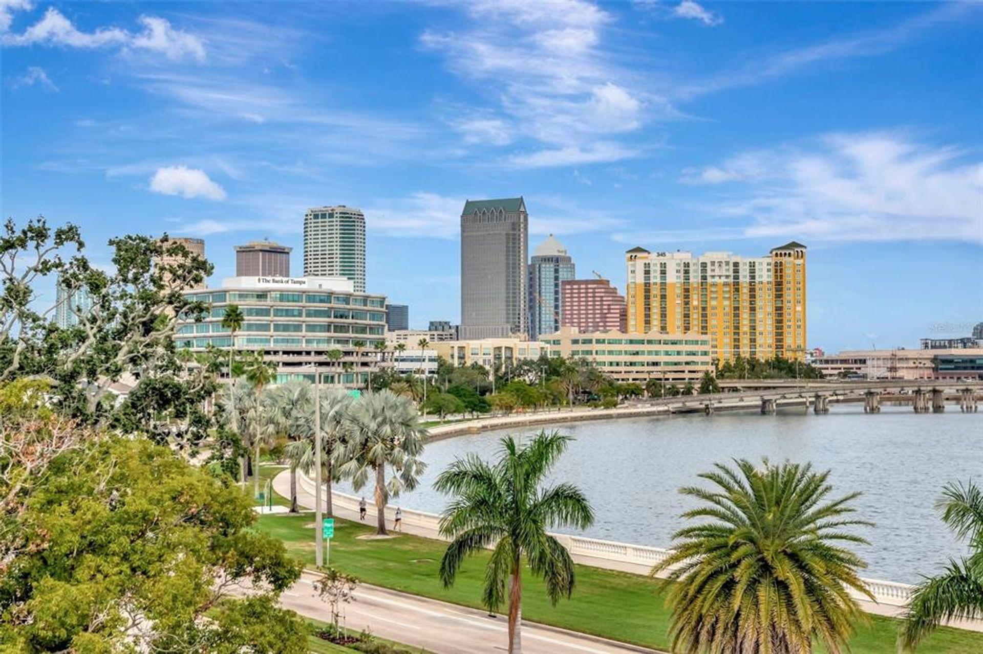 City skyline with tall buildings, trees, a river, and a blue sky with clouds.