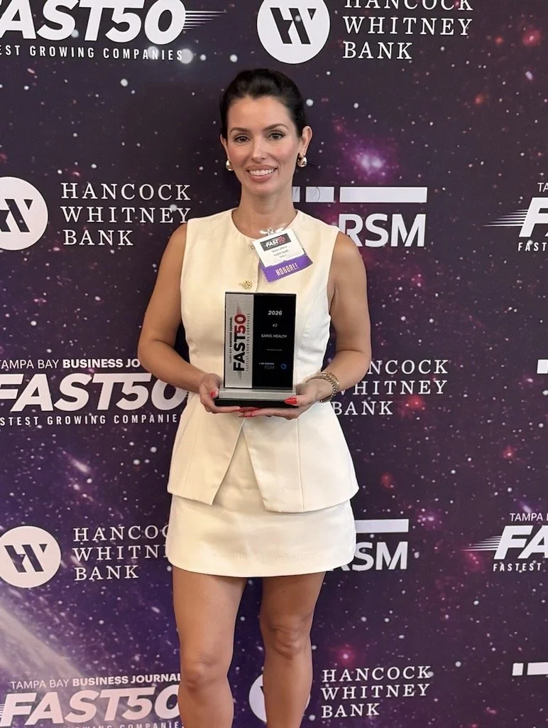 A woman in a cream dress is holding an award plaque at the FAST50 event, with a dark background featuring logos of HANCOCK WHITNEY BANK and FAST50.