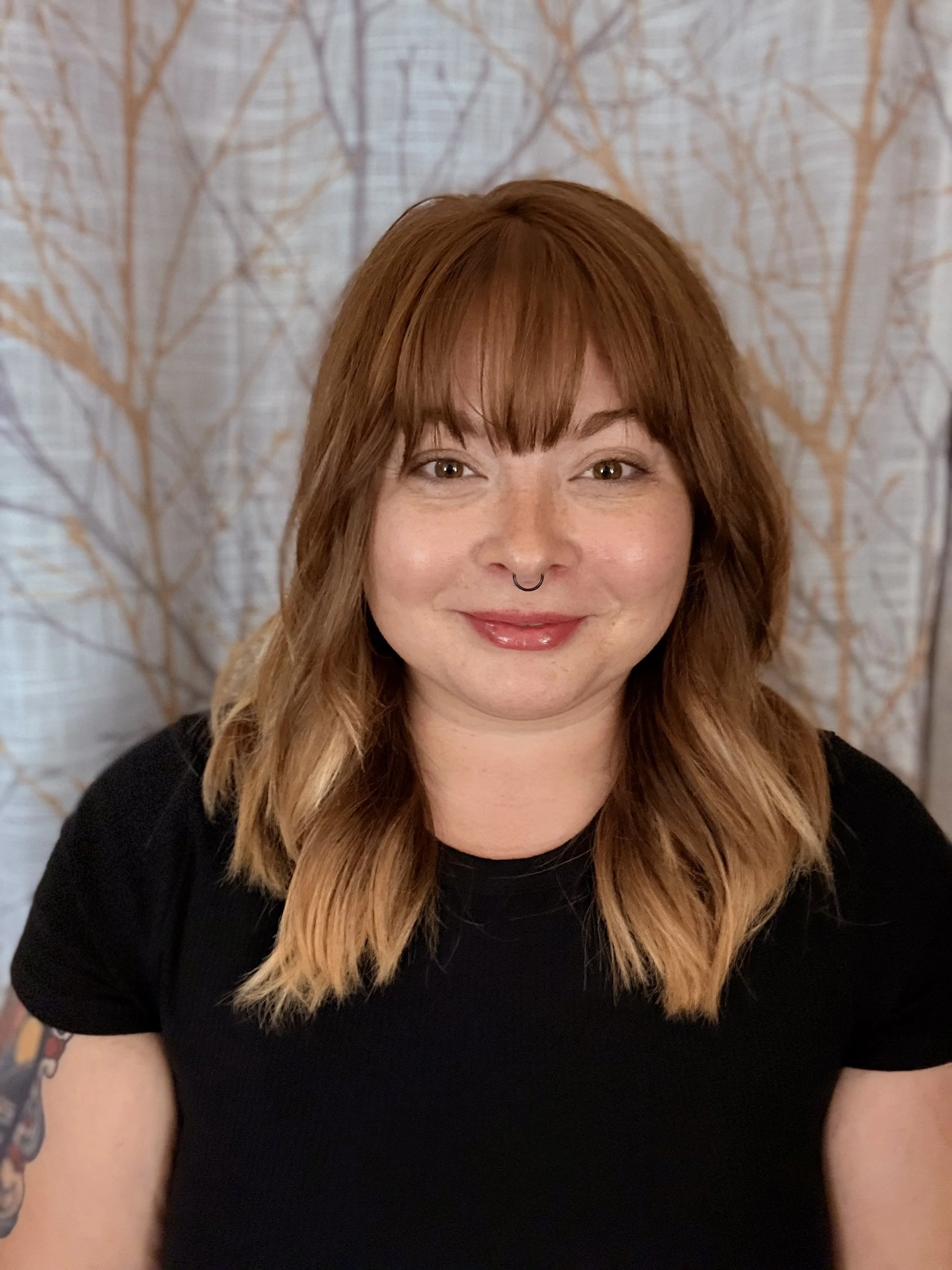 Close-up of a woman with shoulder-length red hair, light skin, and a septum piercing, smiling at the camera, wearing a black top, with a background of patterned curtains featuring a tree branch design.