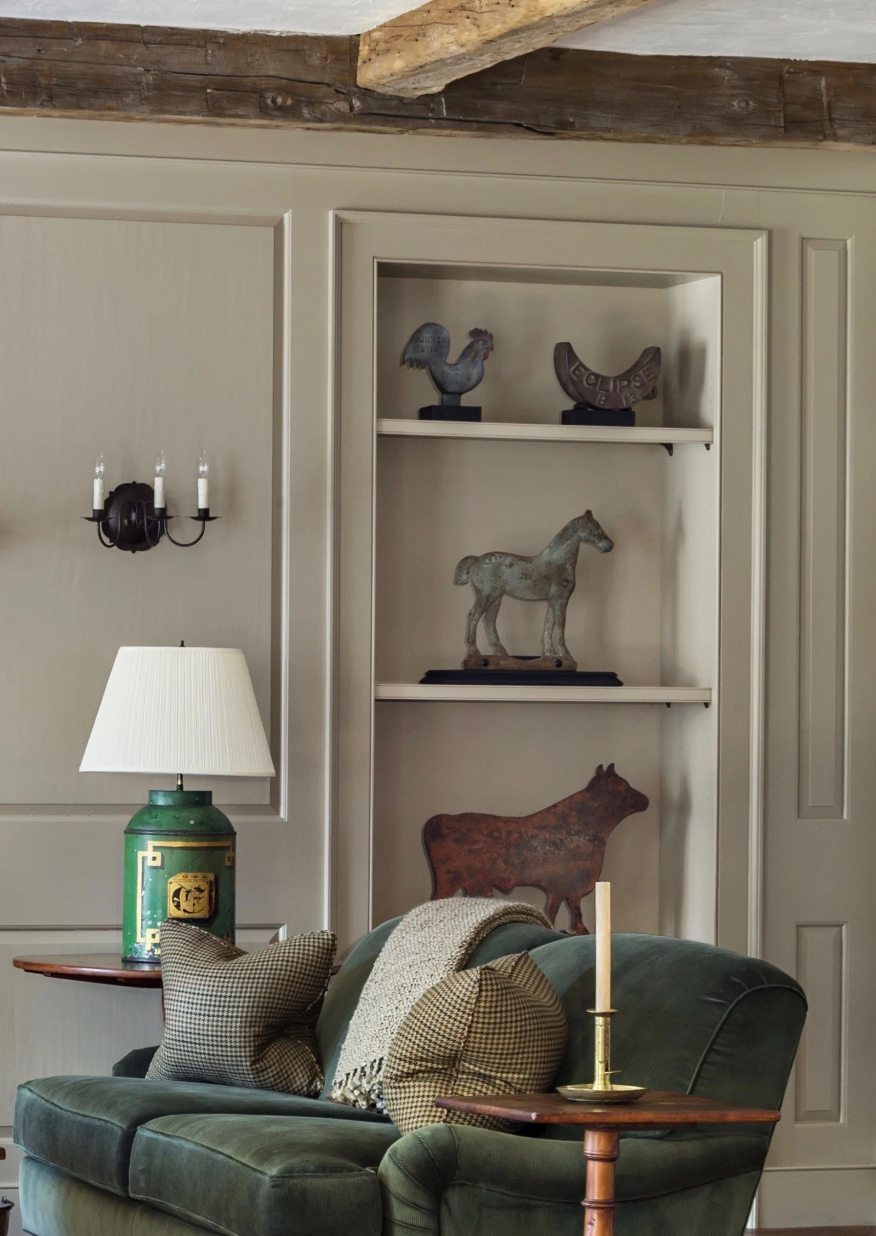 A display of antique 19th century mill weights on shelves within a green paneled wall.   In the foreground is a classic green sofa and table lamp, converted from an antique tea caddy.