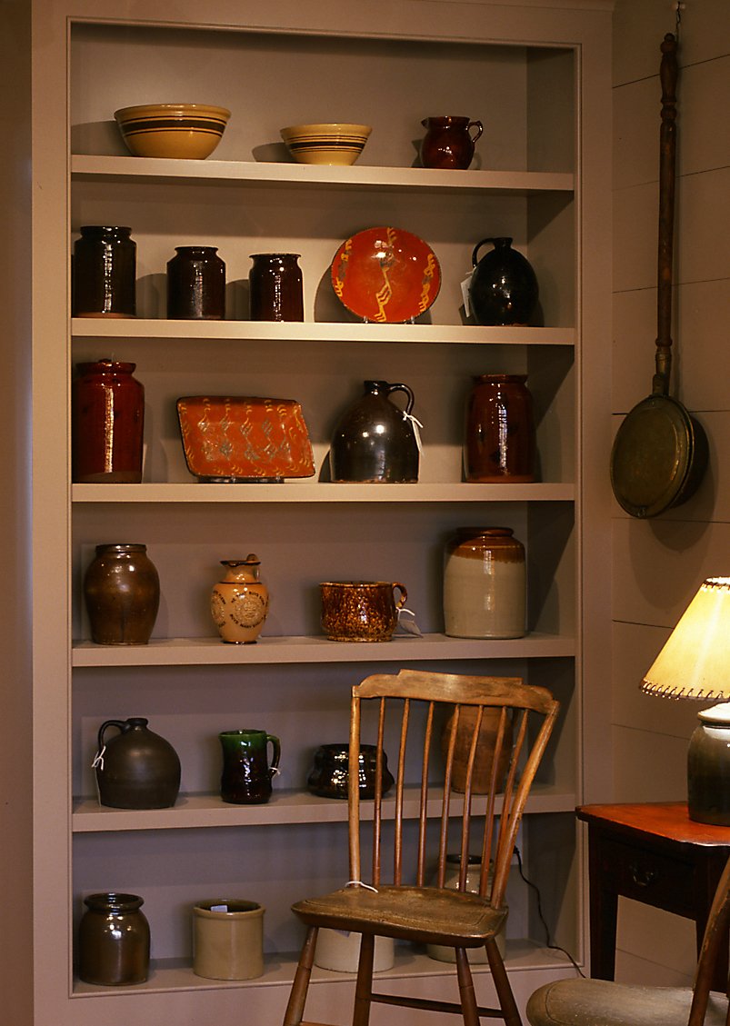 A recessed shelf in an antique shop displaying a collection of American 19th century redware and yellow ware pottery.  A New England step-down Windsor chair and early bed warmer are nearby.