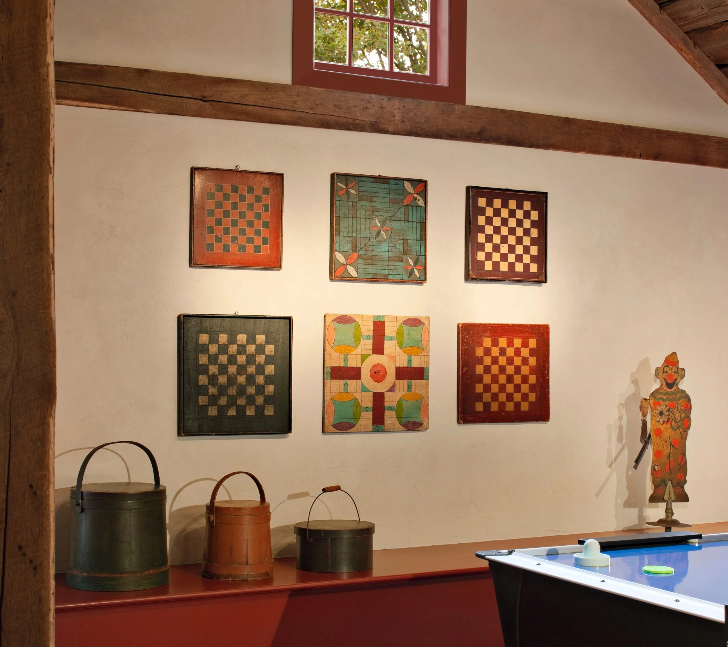 Interior of a converted barn with antique beams.  A collection of antique checker and parcheesi boards is mounted on the wall.  A collection of antique firkins sit on a ledge beside a vintage water sprinkler in the form of a clown
