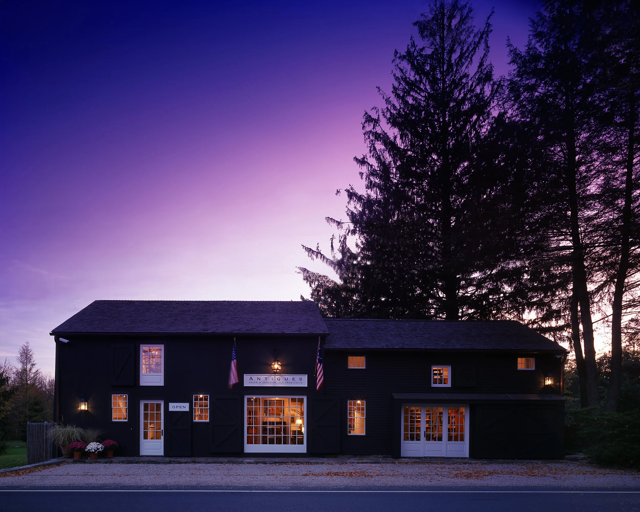 A dark-colored building with large windows and a sign indicating it is an antiques store, with American flags hanging on either side of the entrance. The interior lights are on, showing furniture inside, and there are potted plants with flowers outside. The sky above displays a purple and blue sunset or twilight, with silhouettes of tall trees in the background.