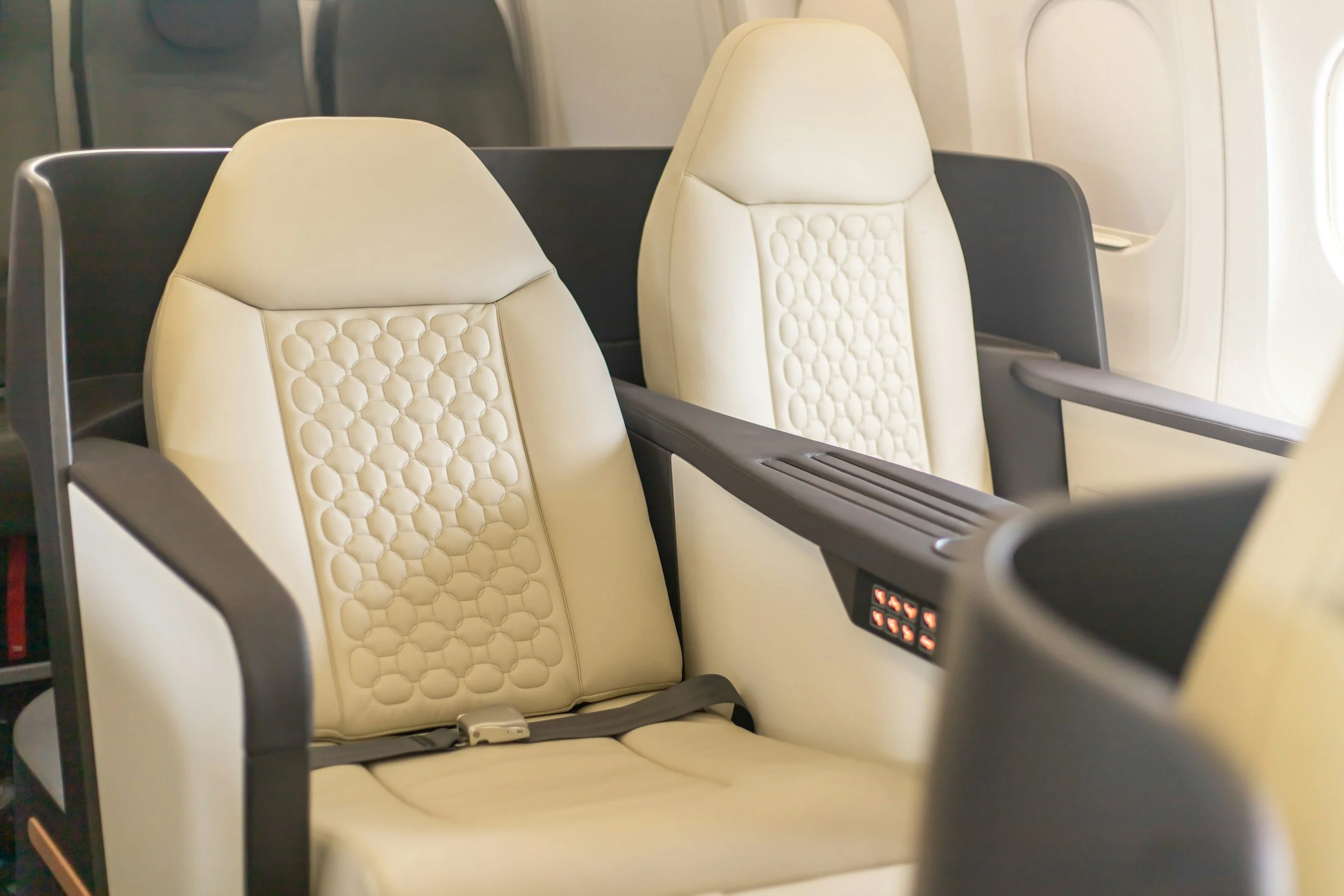 Two beige airplane seats with padded headrests and armrests, located next to a window in an airplane cabin.