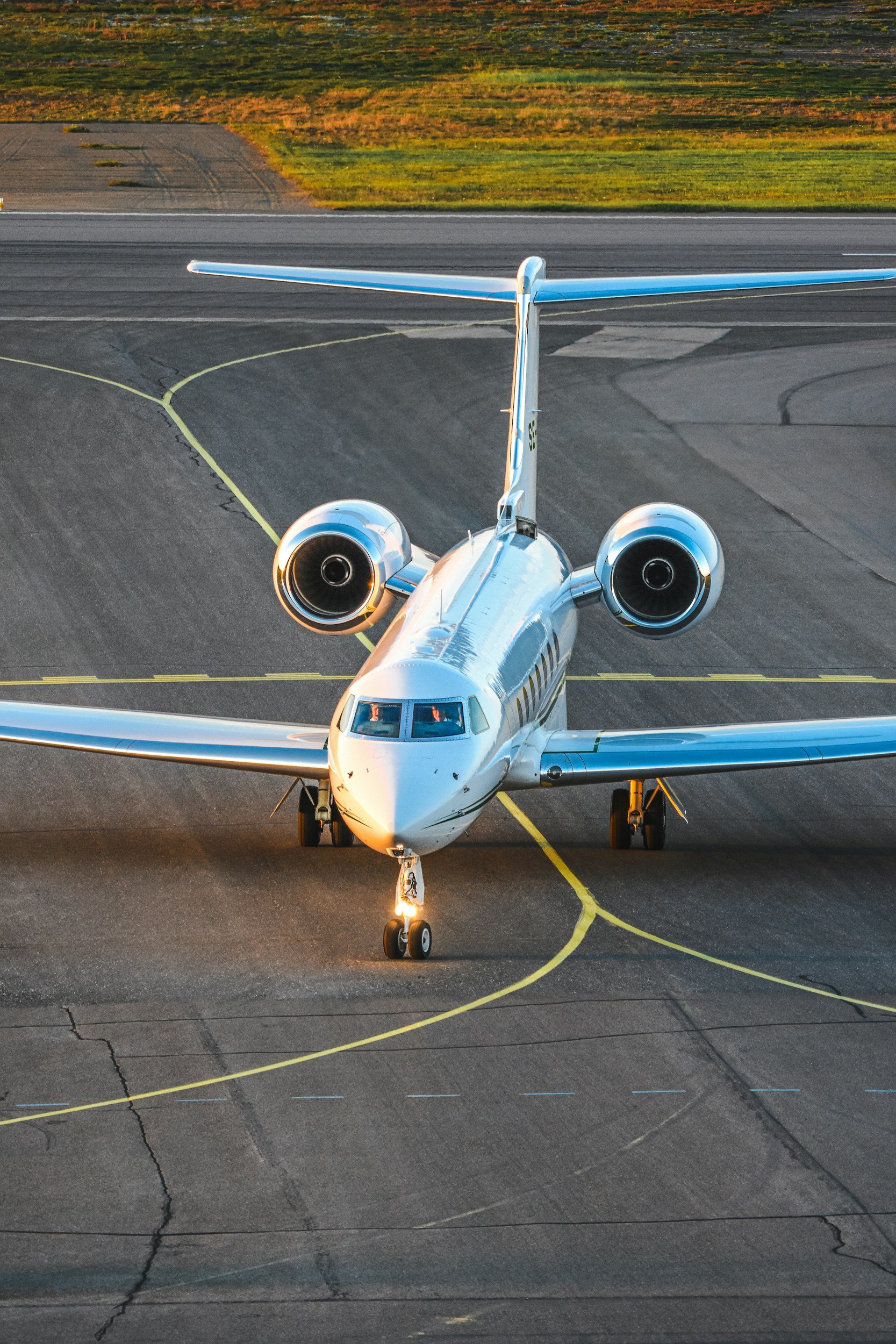 A white private jet aircraft parked on an airport tarmac during sunset, with a grassy field and runway in the background.
