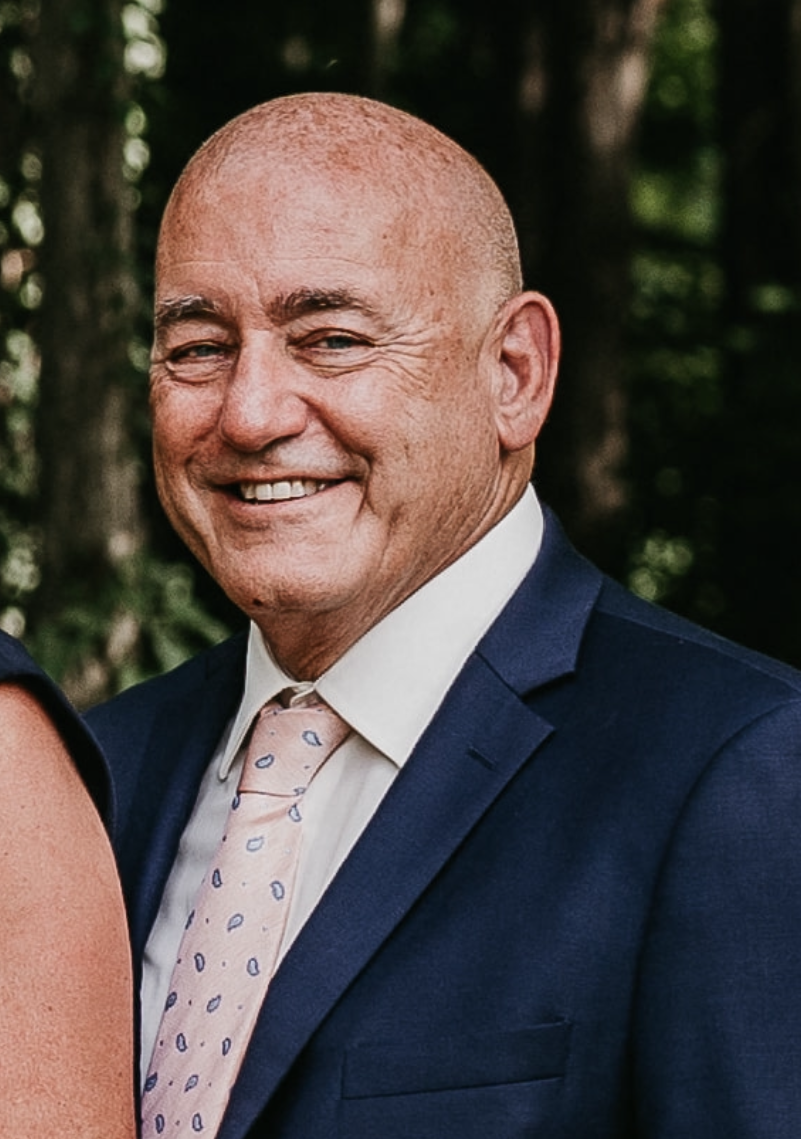 Close-up of a smiling bald man in a formal navy suit, white shirt, and light pink tie with dark blue dots, outdoors with blurred green trees in the background.