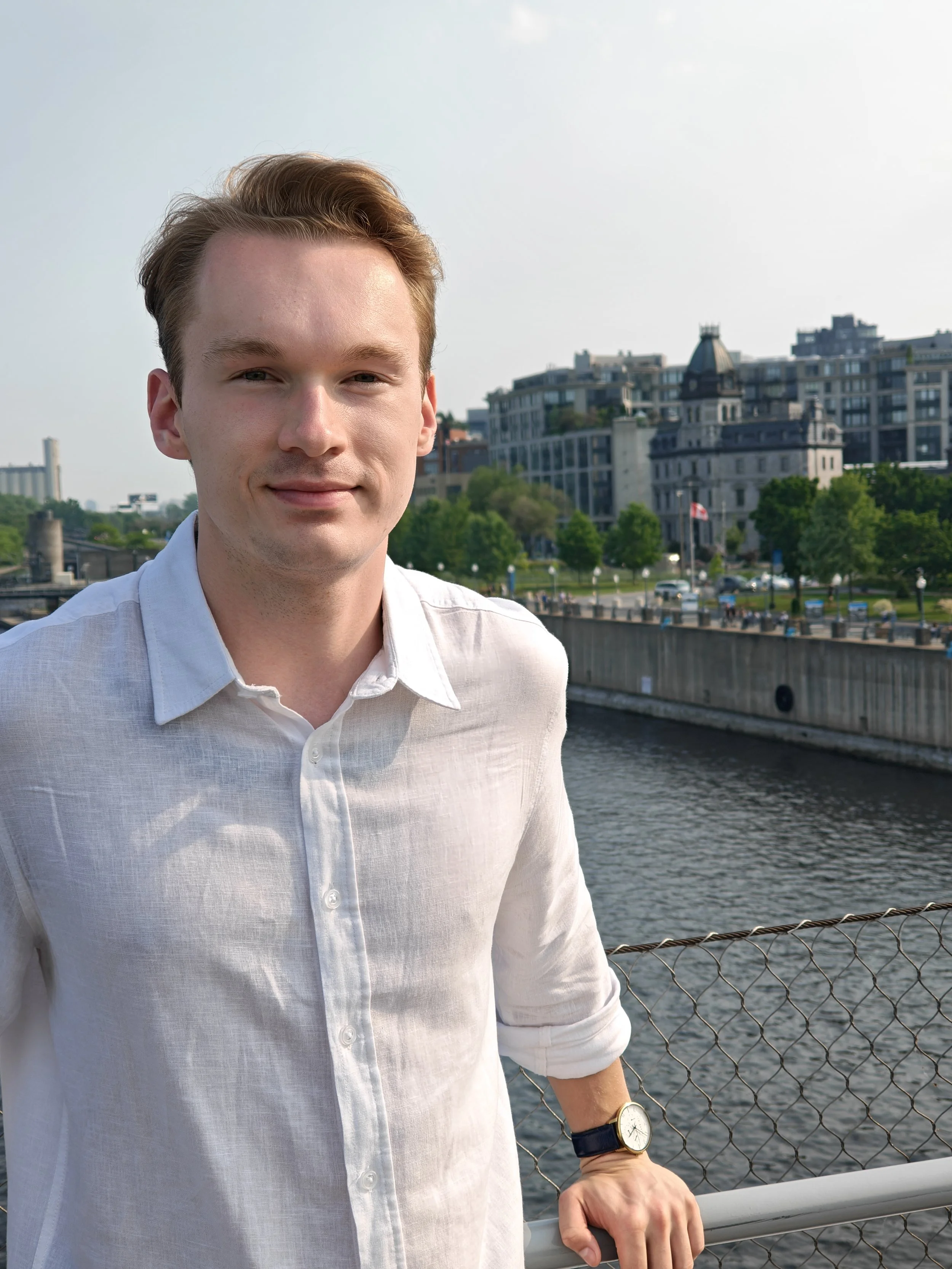 A man in a white shirt standing near a waterway with a cityscape in the background, including buildings and trees, and a Canadian flag visible in the distance.