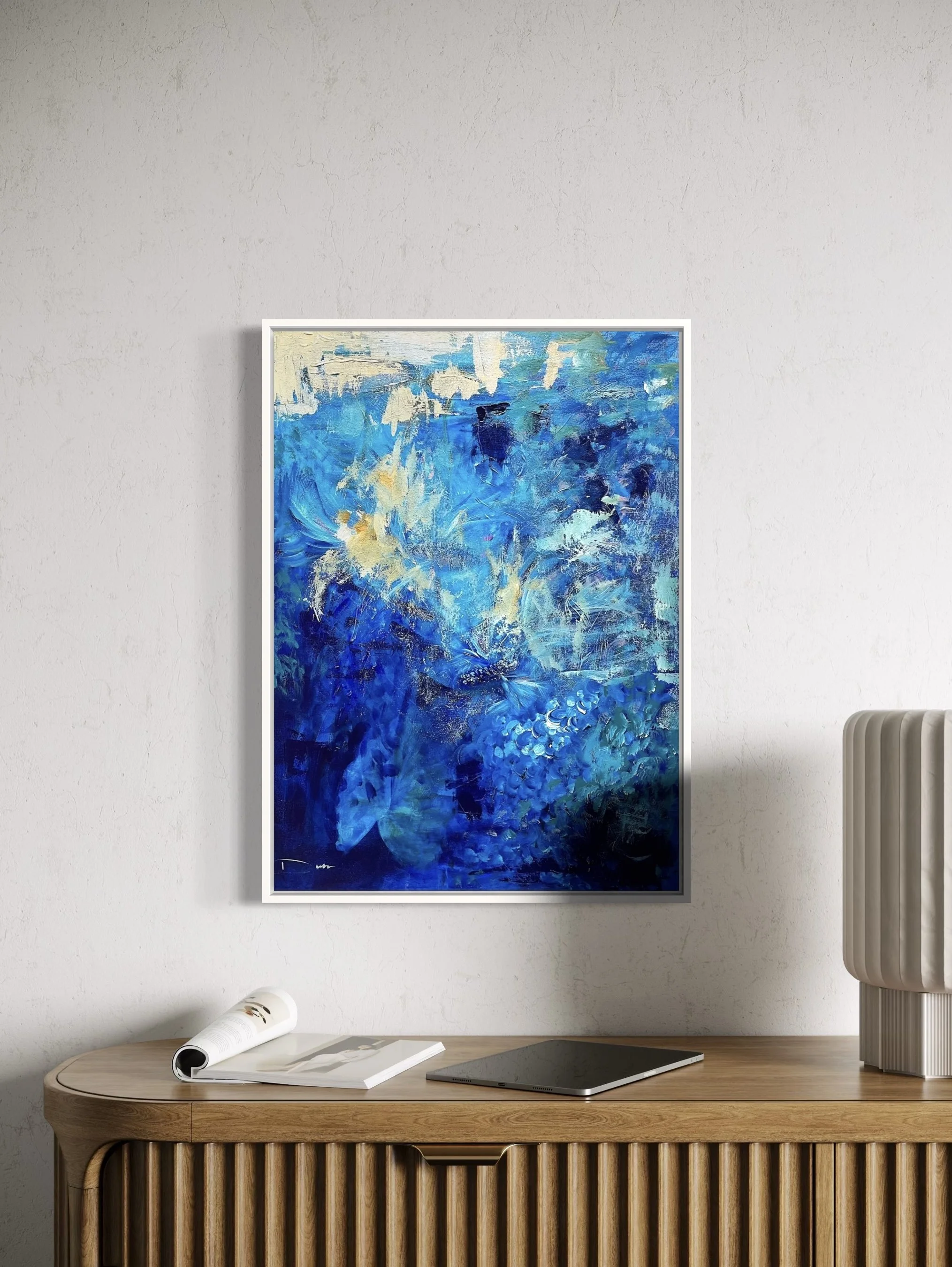Cuadro abstracto con tonos azules, blancos y verdes en una pared blanca. Esquina de un mueble de madera con objetos decorativos, un libro abierto, una laptop y una taza con letras en una mesa de madera.