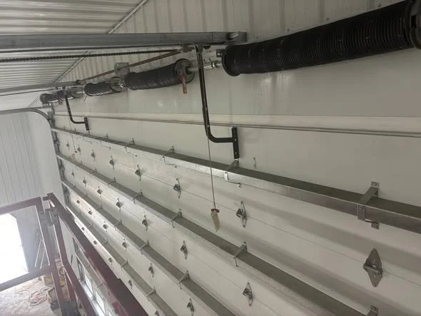 The image shows a large garage door with horizontal panels, equipped with multiple garage door springs along the top. There are black pipes and electrical conduits installed above the door inside a building.