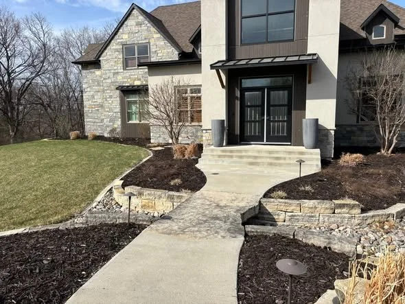 Front entrance of a modern house with concrete steps, black door, and landscaped yard with grass, mulch, and small lights.
