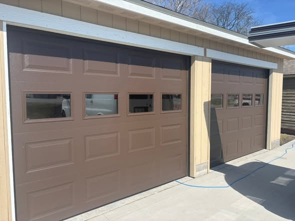 Two brown garage doors with rectangular windows at the top, part of a garage on a house with beige siding.