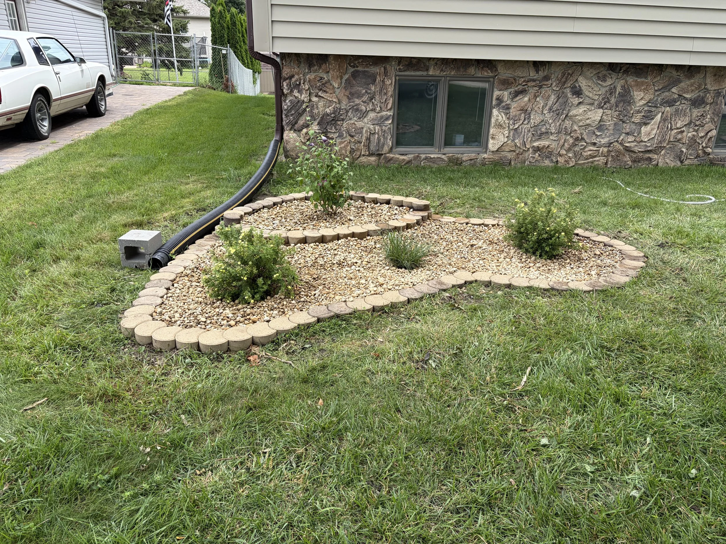 A landscaped yard with a stone border around flower beds, green grass, a window on a stone wall, and a black drainage pipe running along the ground.