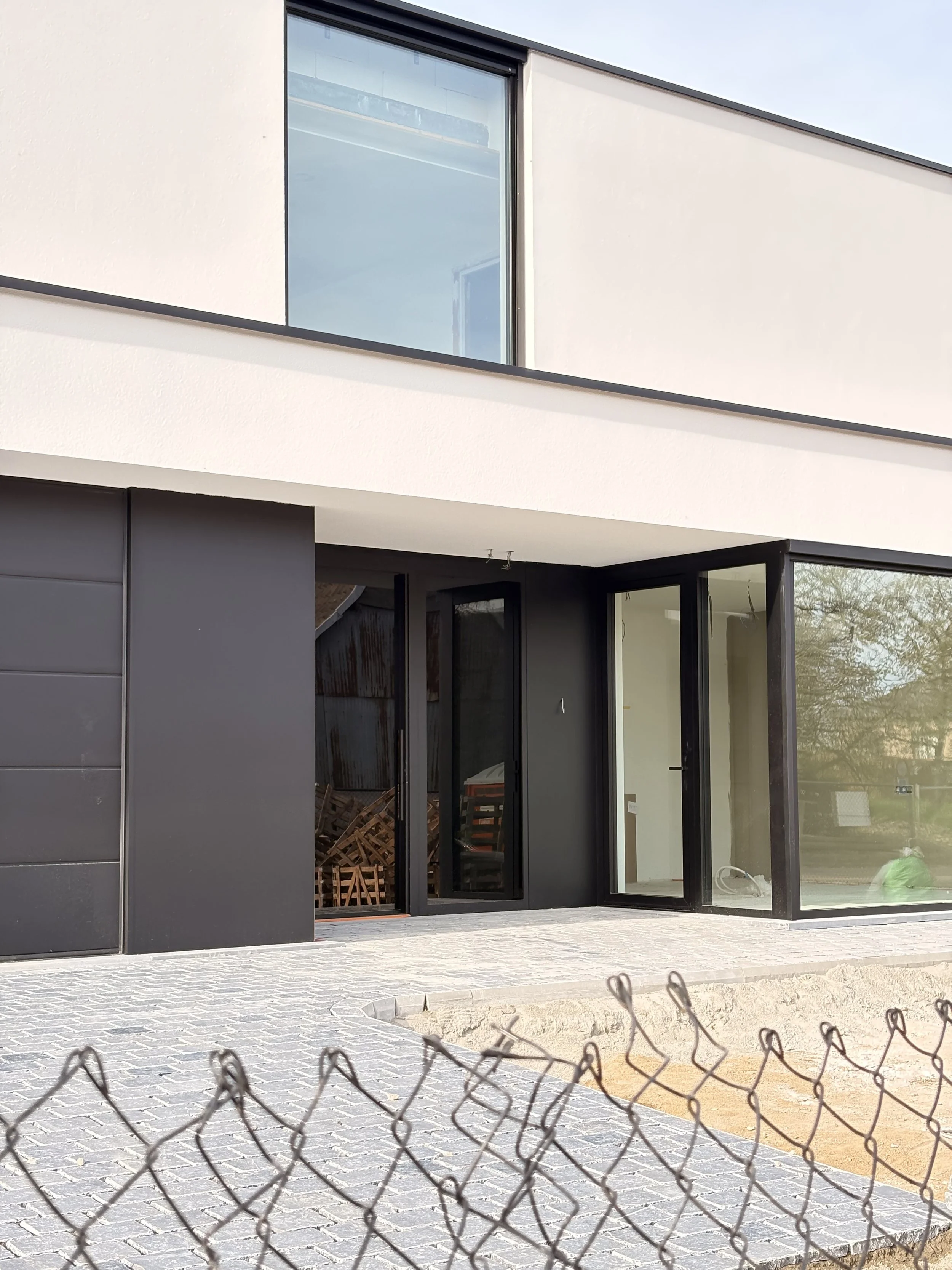 Modern building with large glass sliding doors and white exterior walls, outside paved with cobblestones, surrounded by a chain-link fence.