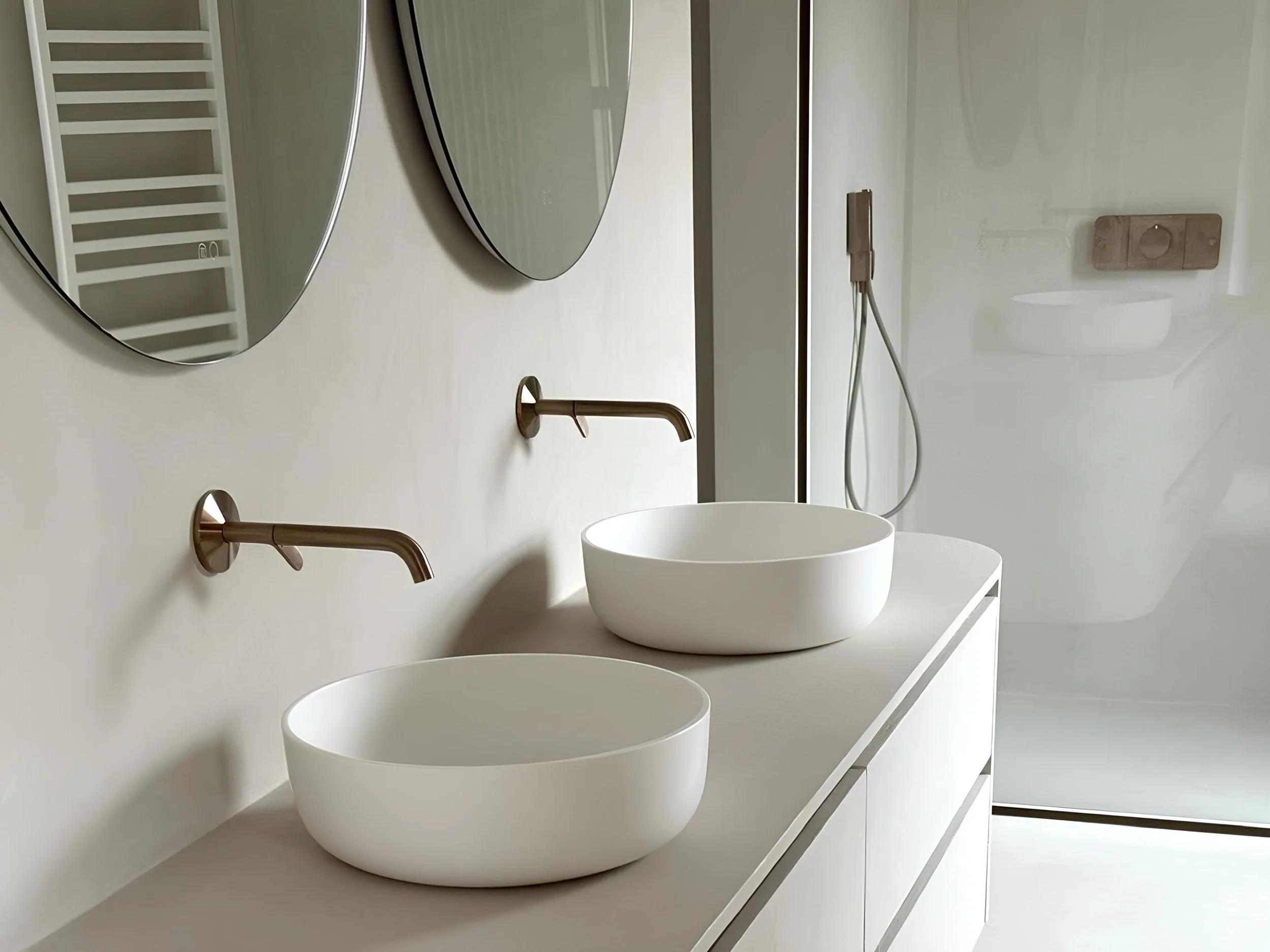 A modern bathroom with three white vessel sinks mounted on a white countertop. Above the sinks are two oval mirrors, and to the left is a white heated towel rack. A brass faucet is mounted between the sinks, and a wall-mounted wooden soap holder is visible on the right side. The wall behind the sinks is white, and there's a power outlet with a plugged-in device on the right.