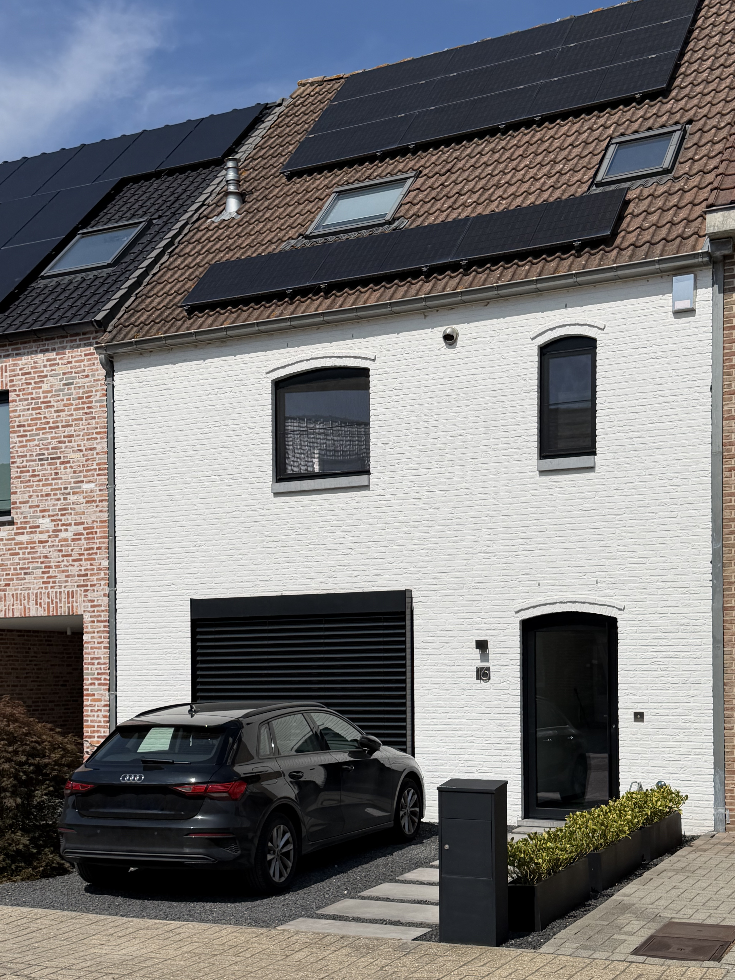 Een modern huis met een witte gevel en zwarte ramen. Een zwarte auto staat geparkeerd voor de woning. Er liggen grind en bestrating voor de ingang, met een tuinbed met planten. Dit is de woning waar MAV Interior Architecture gevestigd is.