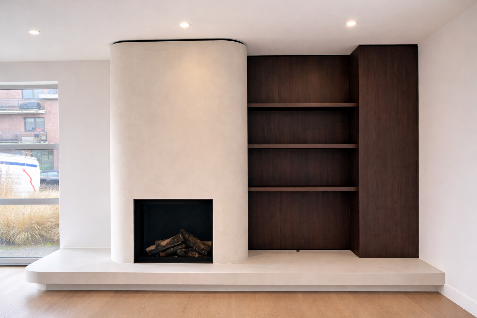 Interior of a modern living room featuring a white curved fireplace with logs inside, next to dark wood built-in shelves on a white wall, with a large window revealing an outdoor urban scene.