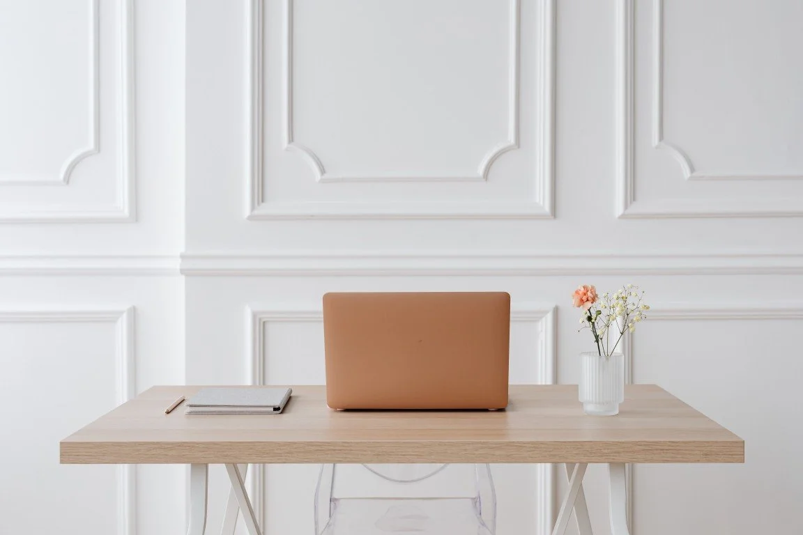 Minimalist wooden desk with a closed copper laptop, a notebook with a pen, and a small vase with pink and white flowers against a white wall with decorative molding.