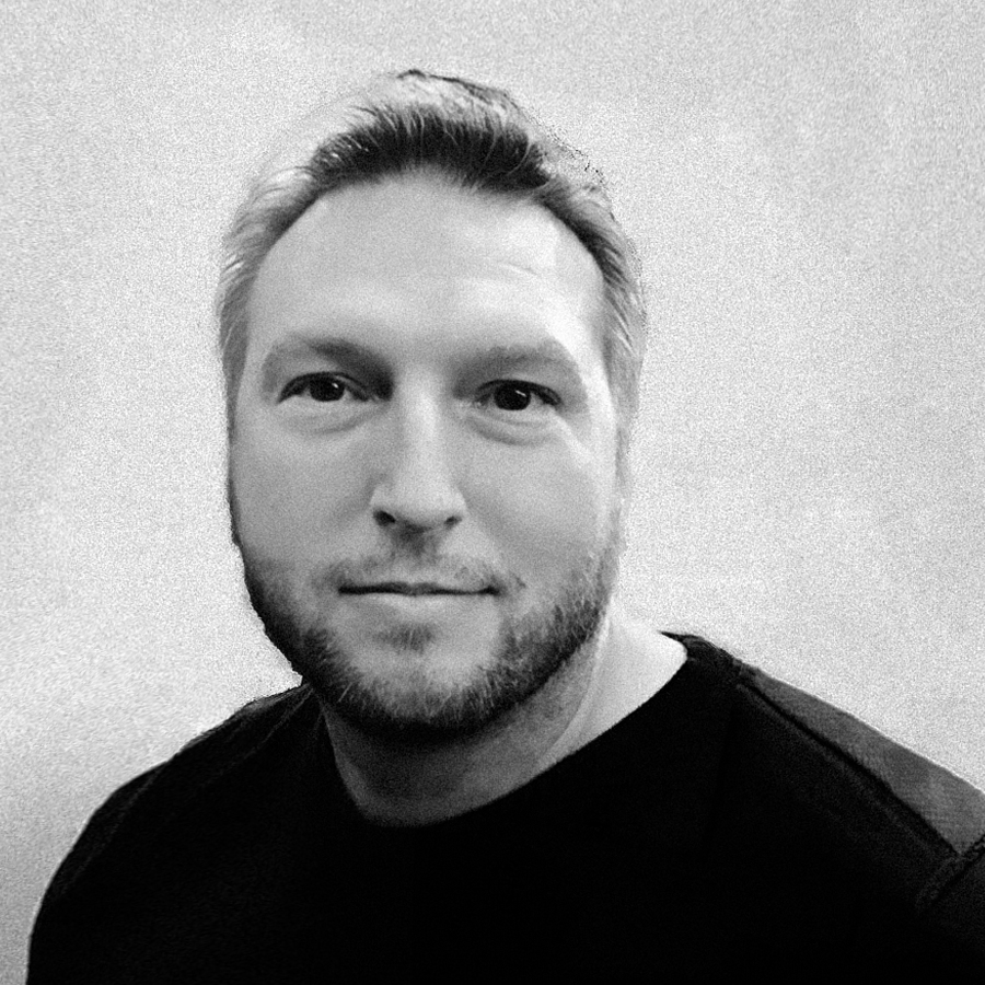 Black and white portrait of a man with short hair and a beard, wearing a dark shirt, smiling slightly, against a plain wall background.