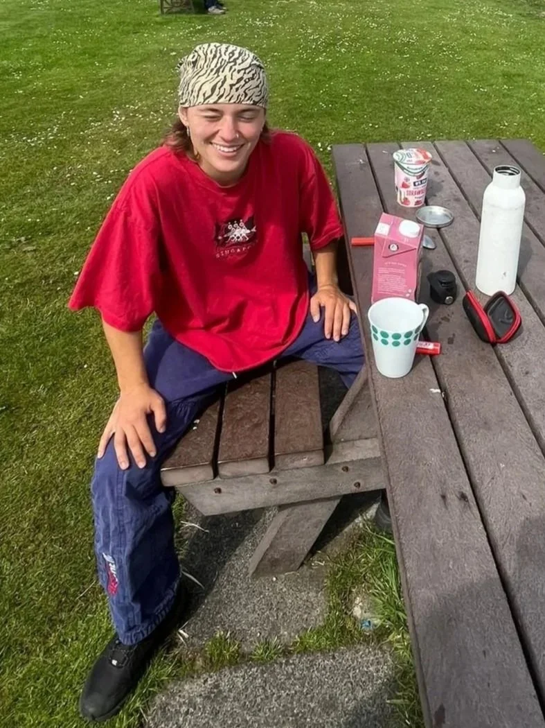 A smiling young person wearing a patterned bandana, red t-shirt, and dark pants sitting on a wooden picnic table outdoors, with a grassy area in the background. On the table are a white water bottle, a mug, a carton of milk, and other small items.