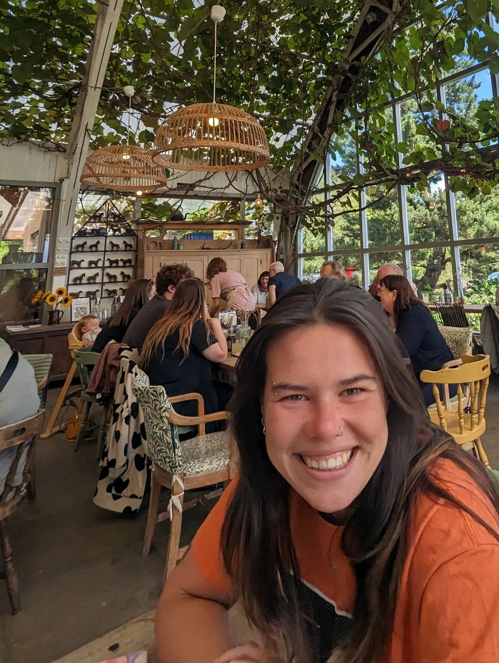A woman with dark hair smiling and leaning over in a restaurant with people sitting at tables, large wicker light fixtures hanging from the ceiling, greenery and large windows showing trees outside.