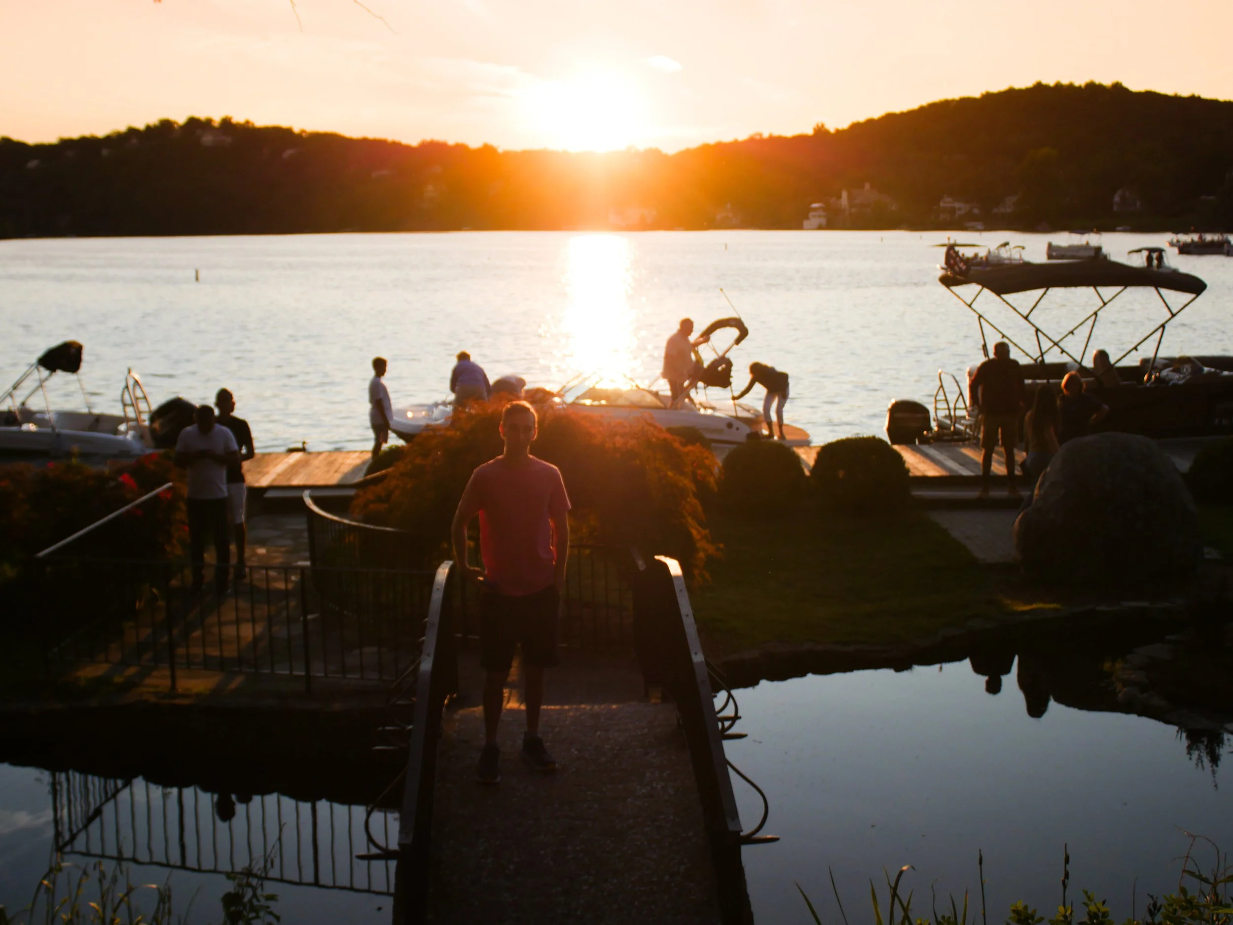 People gather at a dock by the water during sunset with boats in the background, hills across the water, and a reflection of the sunset on the water.