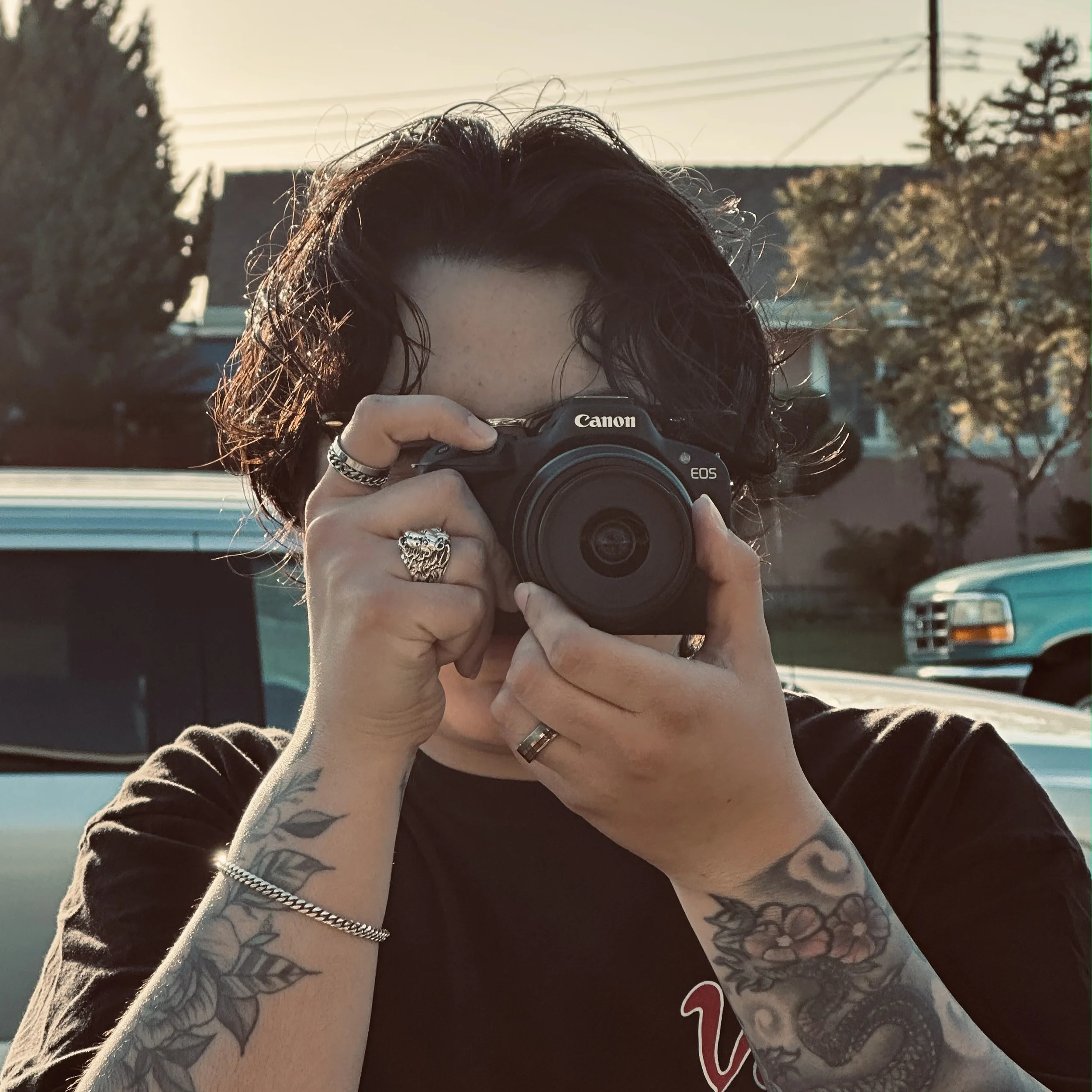 Person with dark curly hair taking a selfie with a Canon camera outdoors with trees, houses, and power lines in the background, wearing a dark shirt and rings on fingers, showing tattoos on arms.