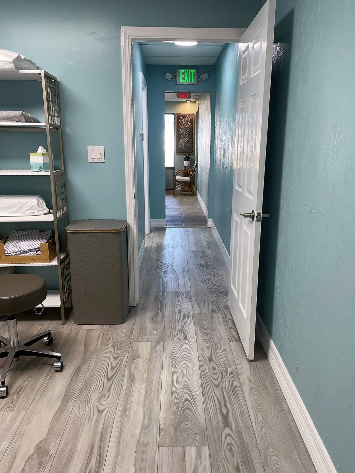 A hallway with blue walls, a white door open to the right, and green 'EXIT' and red 'EXIT' signs above the hallway. To the left, there are shelves with bedding and a grey tissue box. The floor has light wood grain texture.