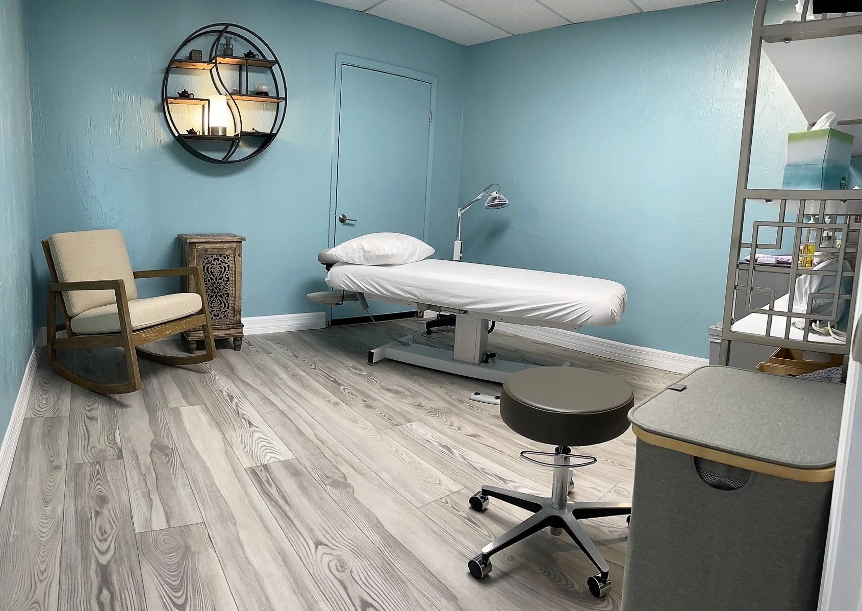 A medical examination room with light blue walls, a white adjustable examination table with a pillow, a small wooden side table, a beige armchair, a round stool, a metal shelving unit with medical supplies, and a decorative wall shelf with small decor items and a lamp.