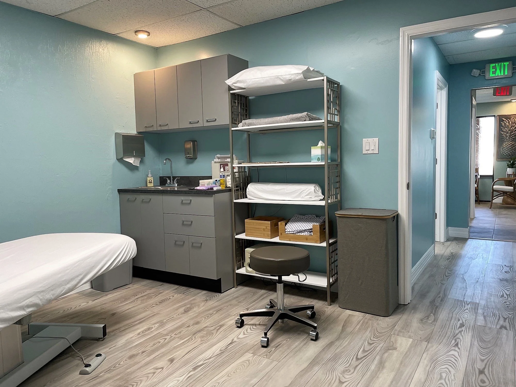 Hospital room with a bed on the left, a cabinet with medical supplies on the right, and a rolling stool in the middle. There is a metal shelving unit with sheets and boxes, and a small sink area with soap and paper towel dispenser. The room has light blue walls and wood-textured flooring, with an open door leading to a corridor with exit signs.