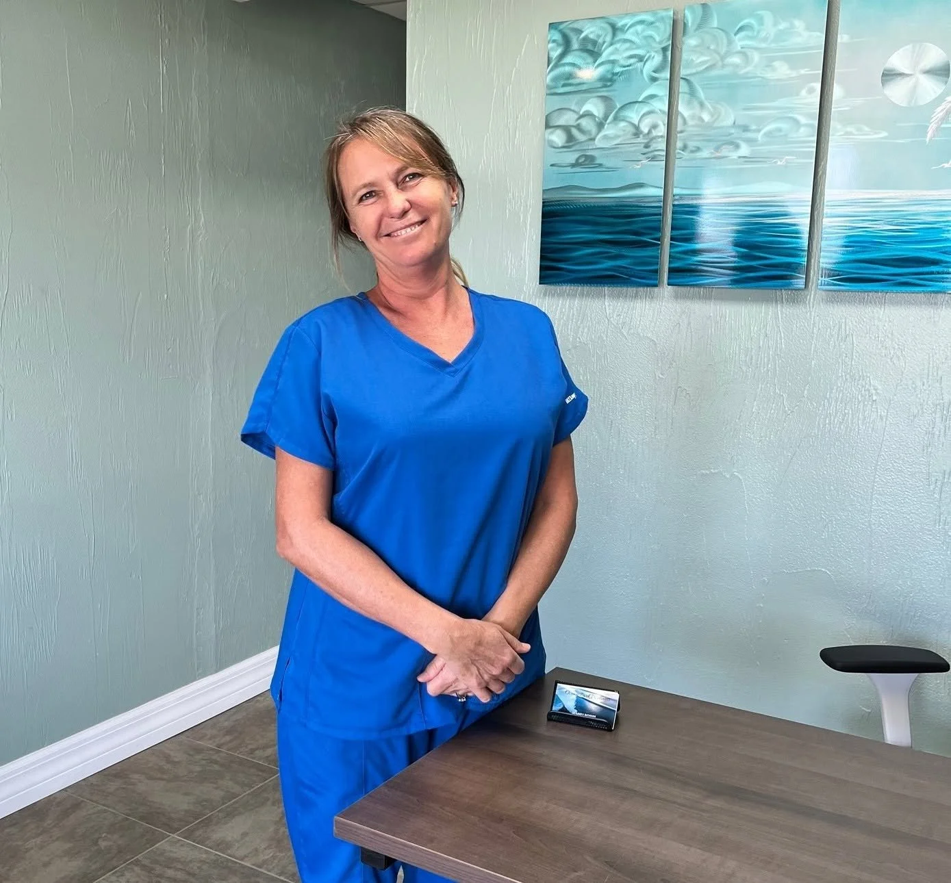 Dr. Lanika in blue medical scrubs standing in a room with a green textured wall, a multi-panel ocean-themed painting, and a wooden table with a small electronic device on it.